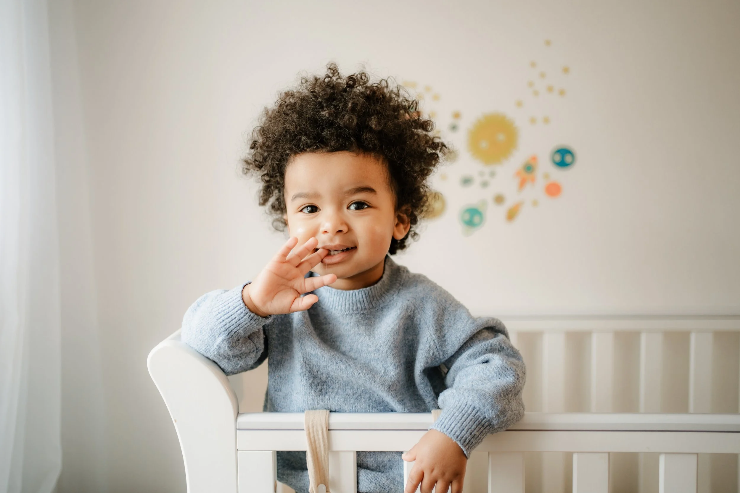 A young child with curly hair leaning on a white crib, wearing a gray sweater, holding his hand near his mouth, in a room with cream-colored walls decorated with colorful abstract shapes.