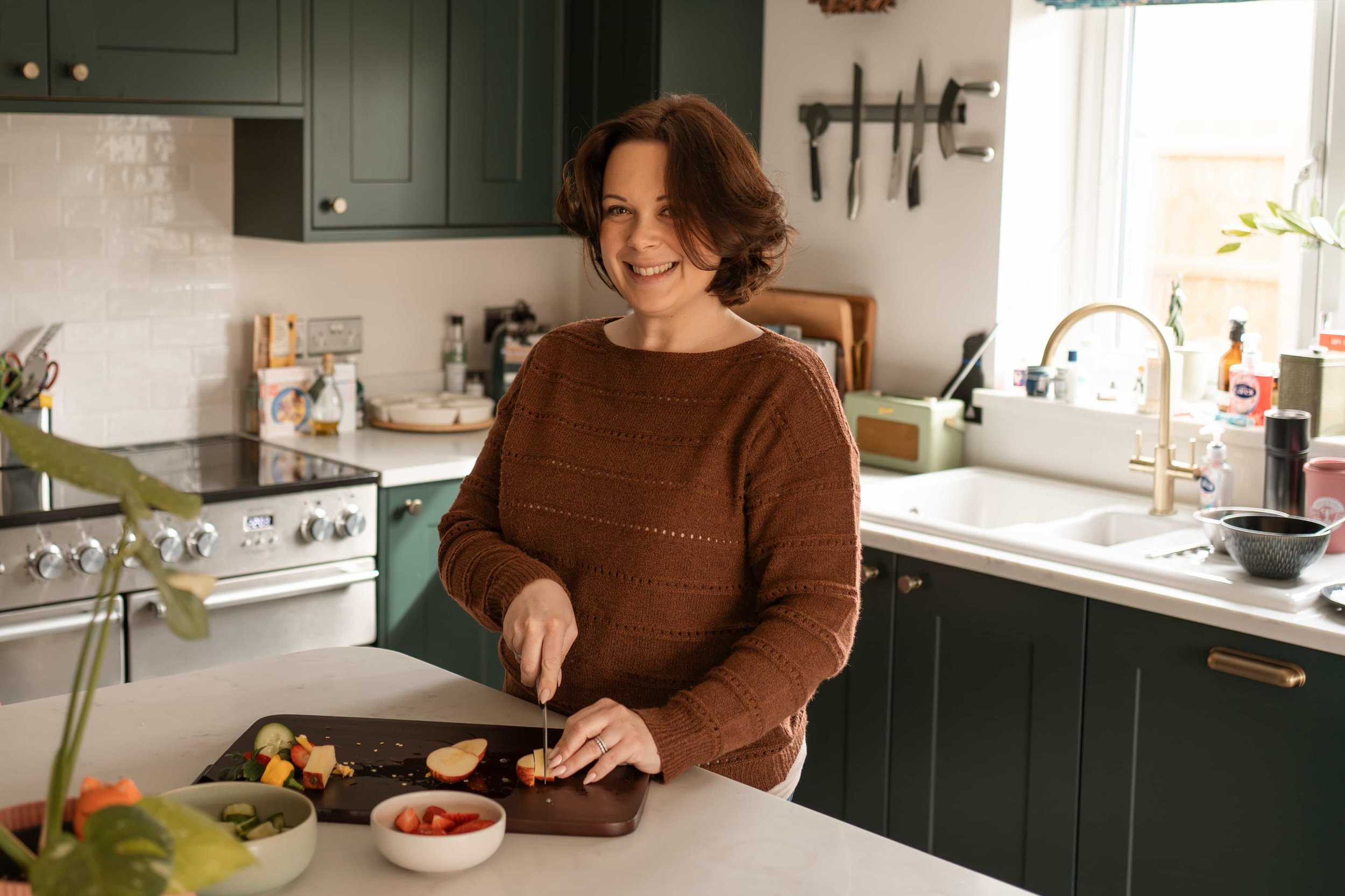 A woman with short curly brown hair smiling while slicing fruit on a black cutting board in a kitchen with green cabinets and a white countertop, with various bowls of food and kitchen utensils visible in the background.