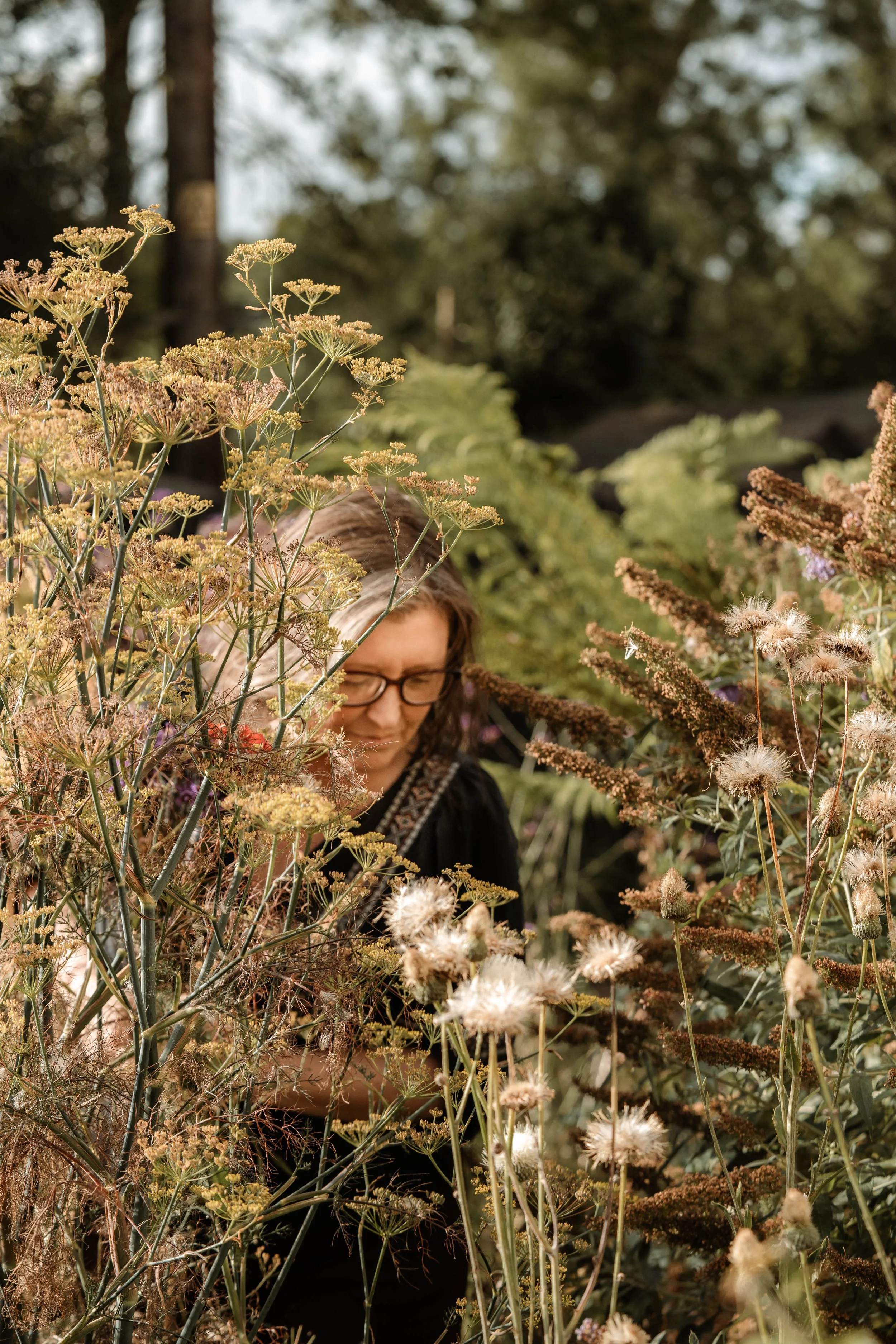 A woman with glasses and dark hair in a garden, surrounded by various plants and flowers.