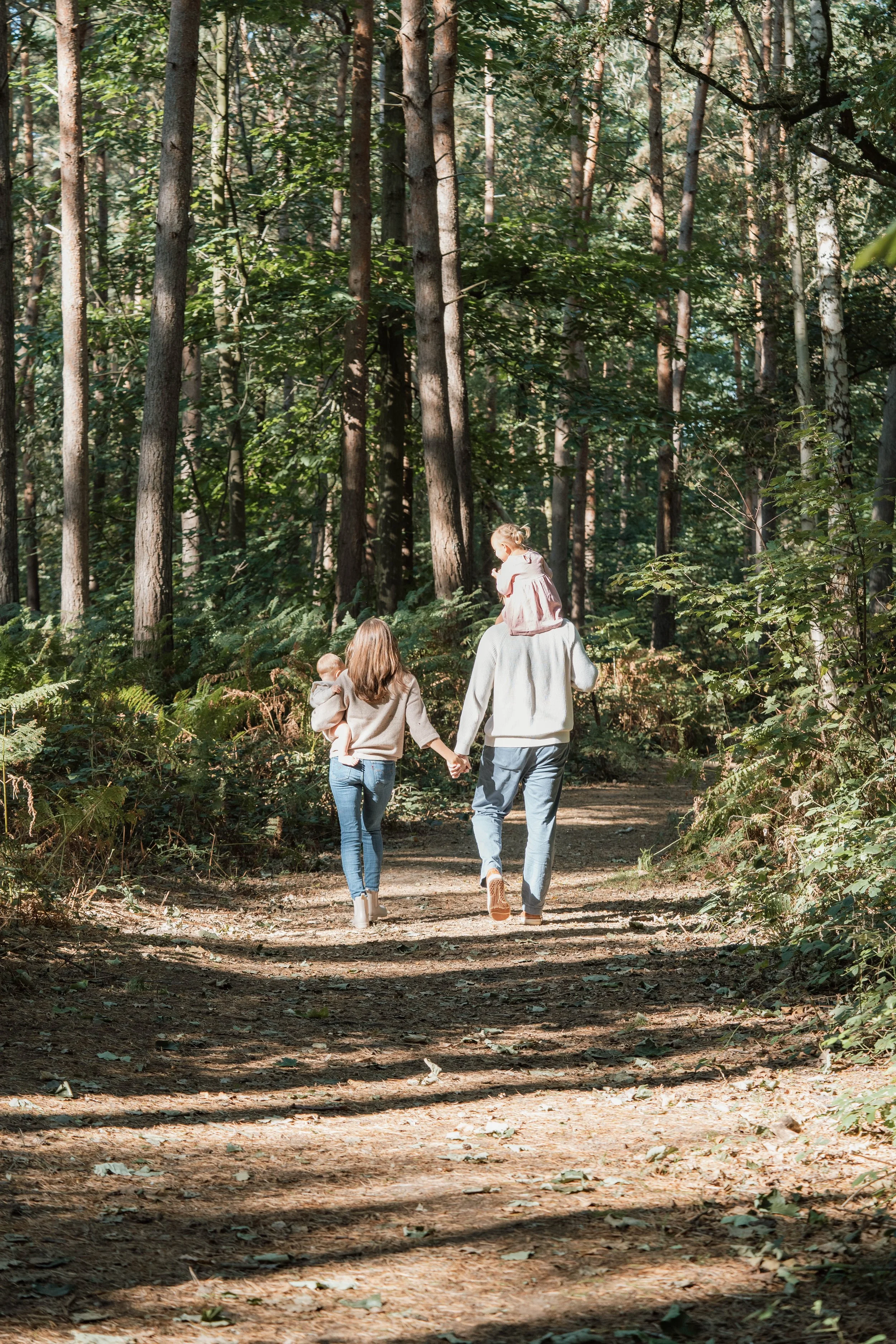 A family of four walking on a forest trail, holding hands, with a woman and a man carrying two children, surrounded by tall trees and green foliage.