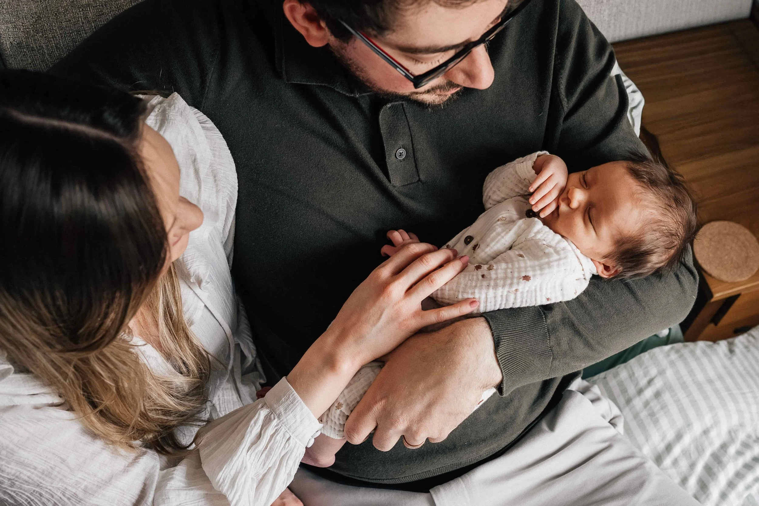 A man holding a sleeping baby on his lap, with a woman gently touching the baby's hand.