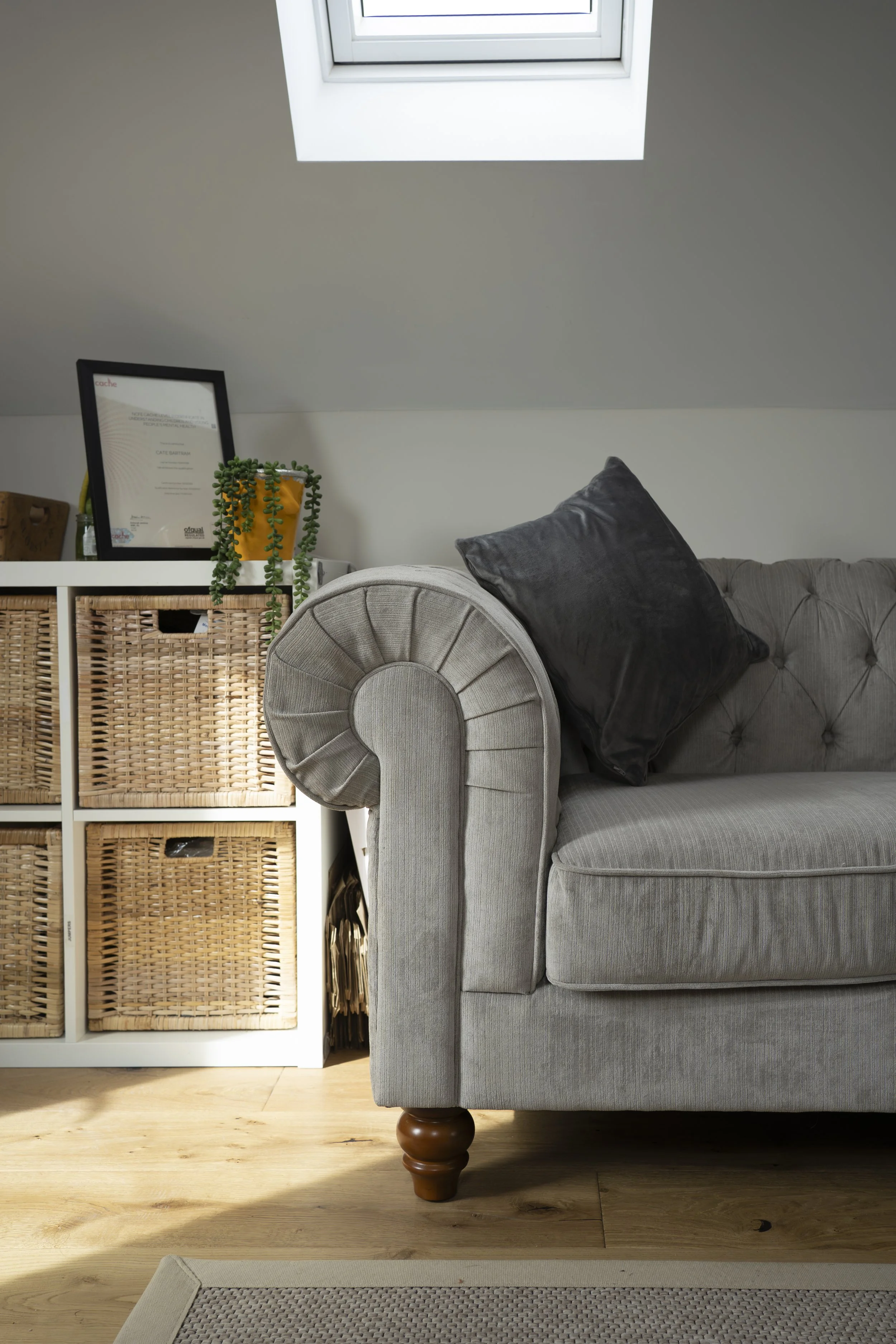 Close-up of a gray upholstered sofa with a black velvet pillow, situated on a wooden floor in a room with a sloped ceiling and a skylight window. To the left is a white shelving unit with wicker baskets and a framed certificate, and a small potted pl
