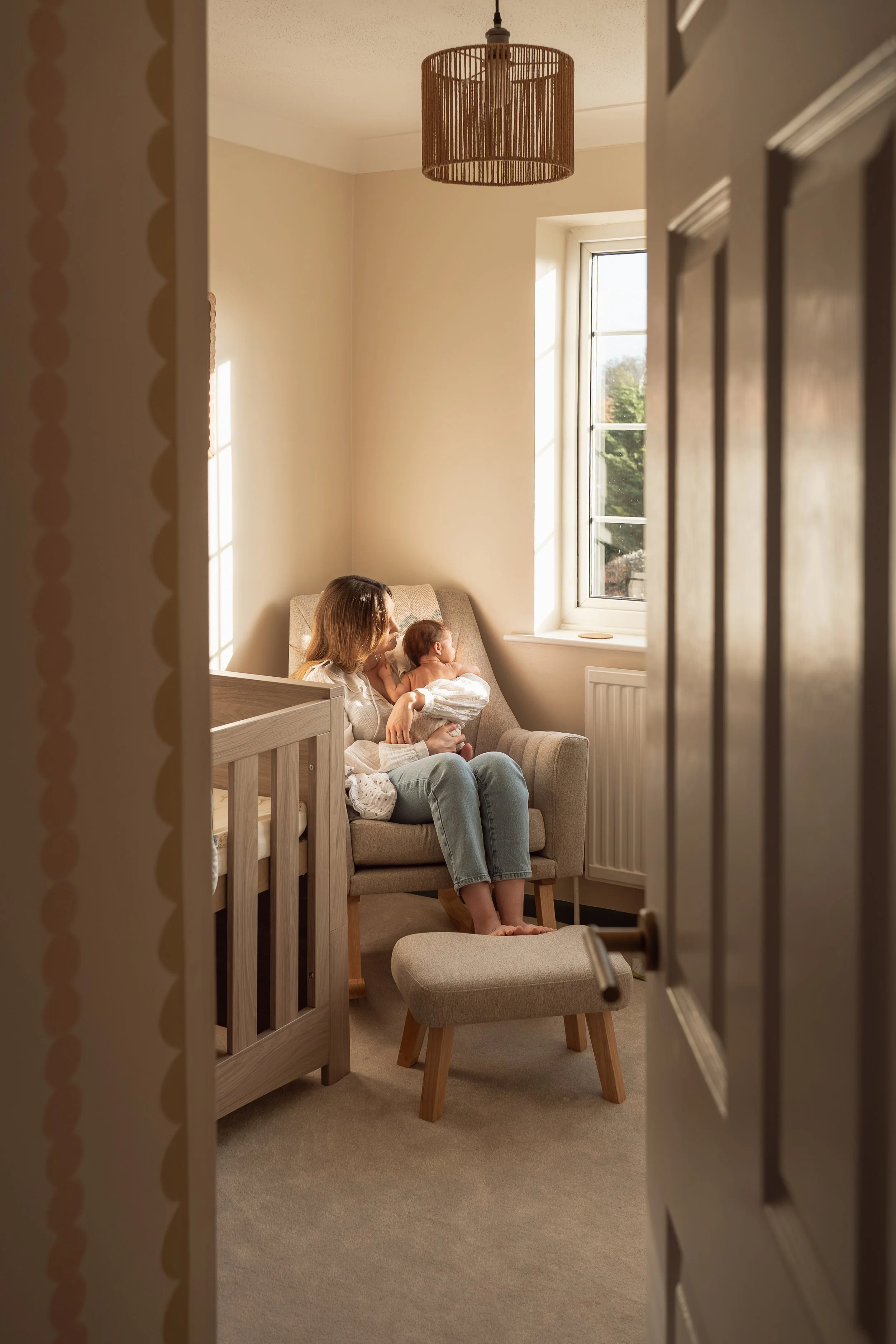 A woman sitting on an armchair with a baby on her lap, seen through a partially open door in a brightly lit nursery with a window, a crib, and a small footstool.