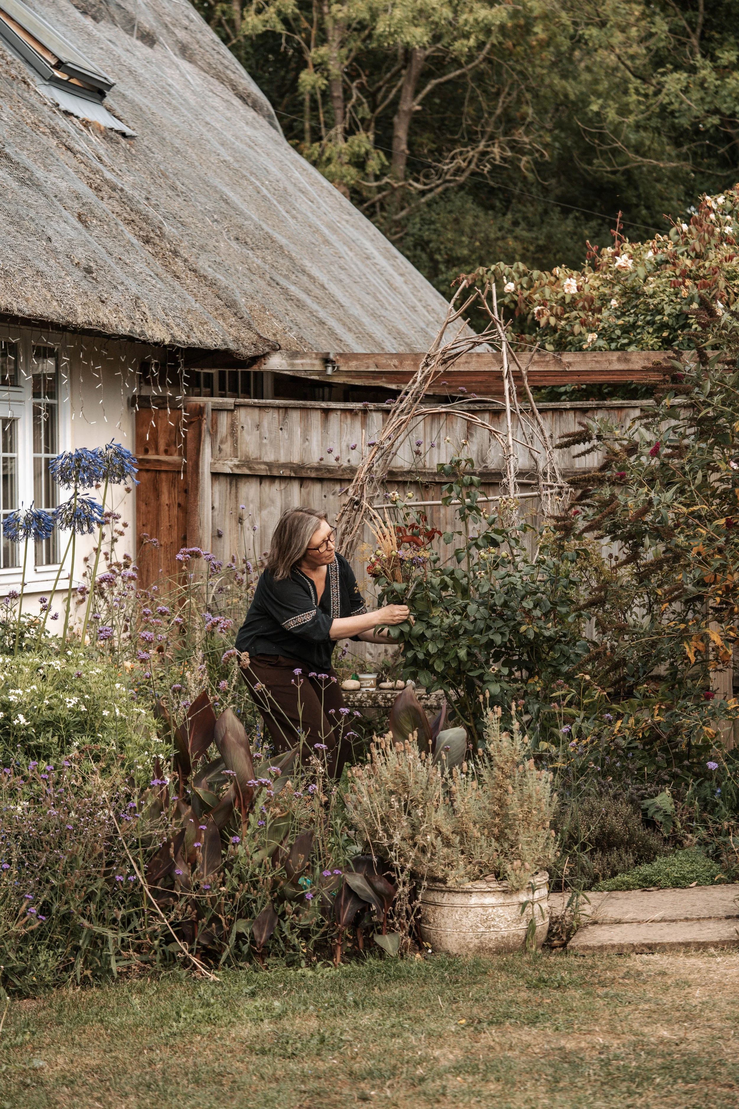 A woman tending to a garden behind a wooden fence, with a thatched roof house in the background.