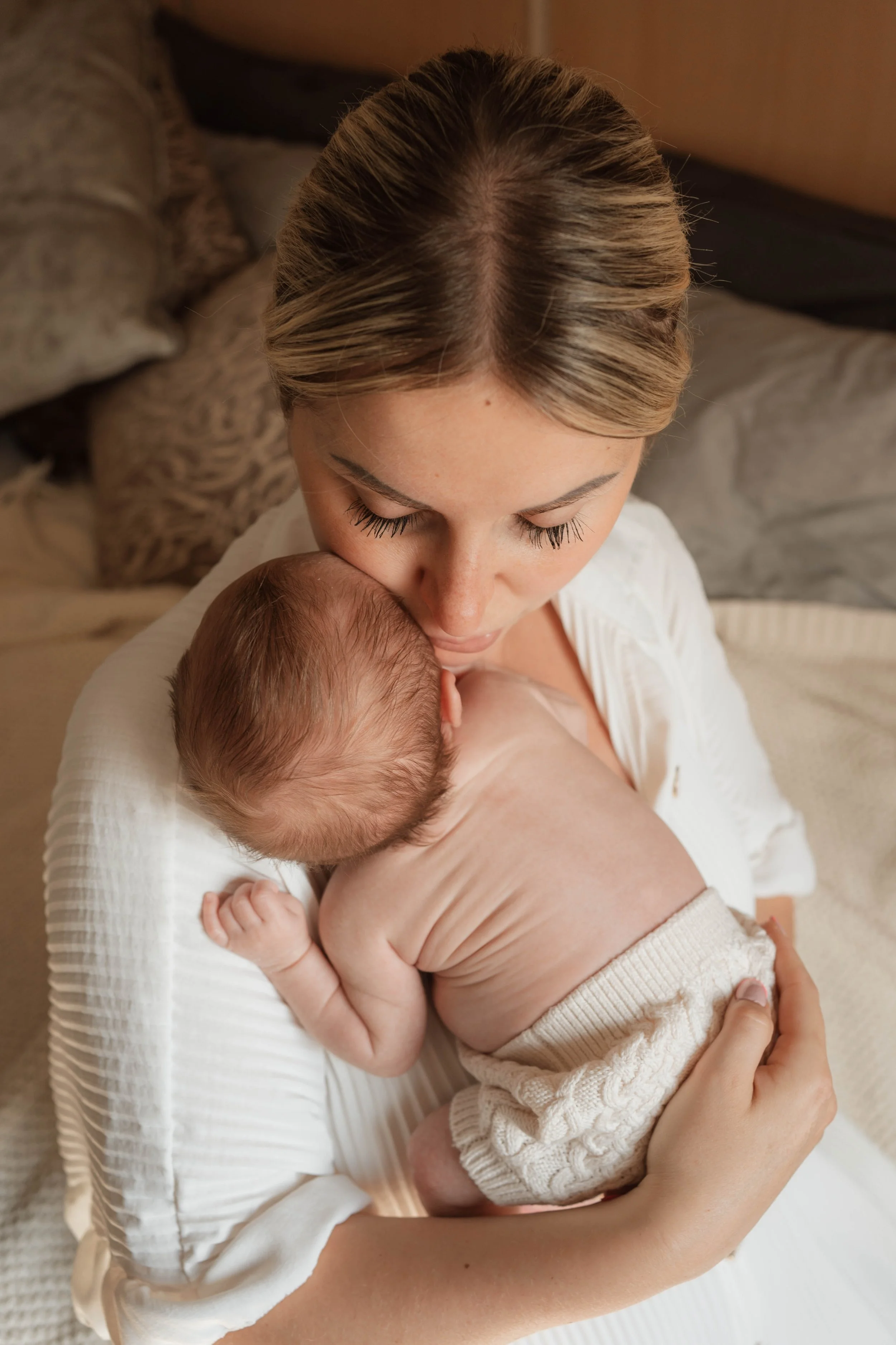 A woman with blonde hair cradles a newborn baby, holding the baby close to her chest while sitting on a bed in a cozy bedroom.