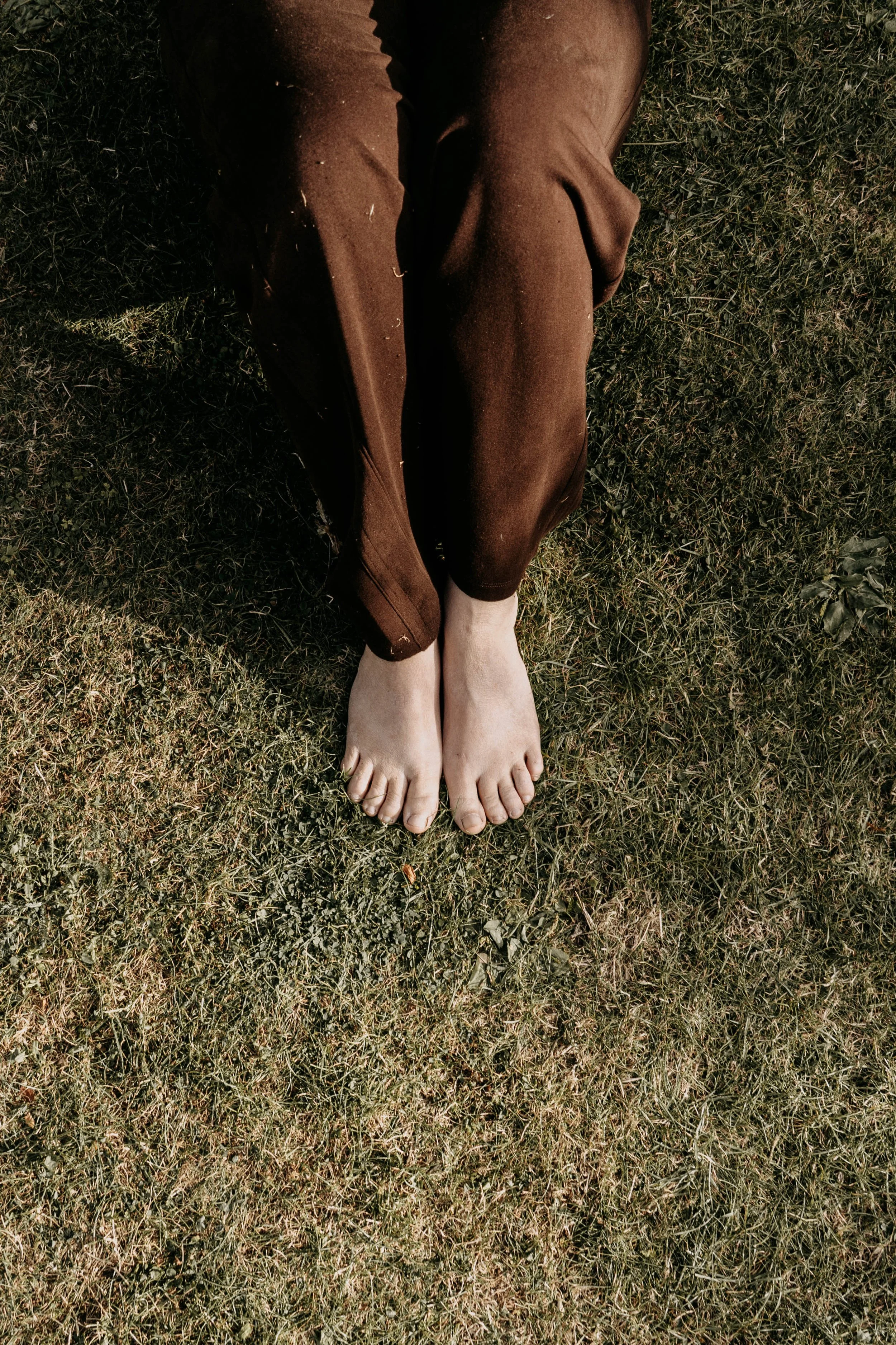 Person sitting outdoors on grass with legs stretched out, wearing brown pants and barefoot.