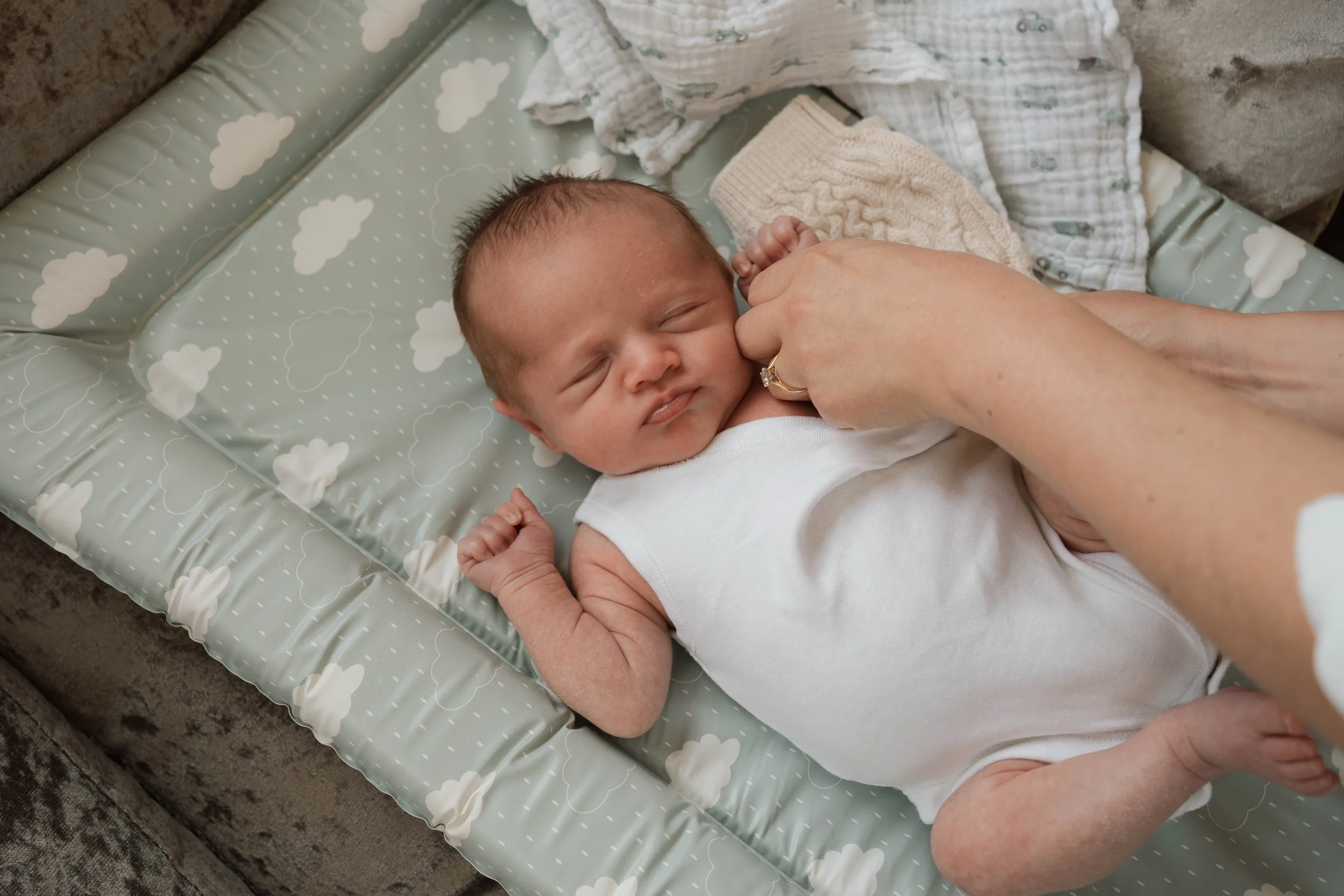 A newborn baby lying on a cushioned changing pad with cloud patterns, being gently touched and comforted by a woman.