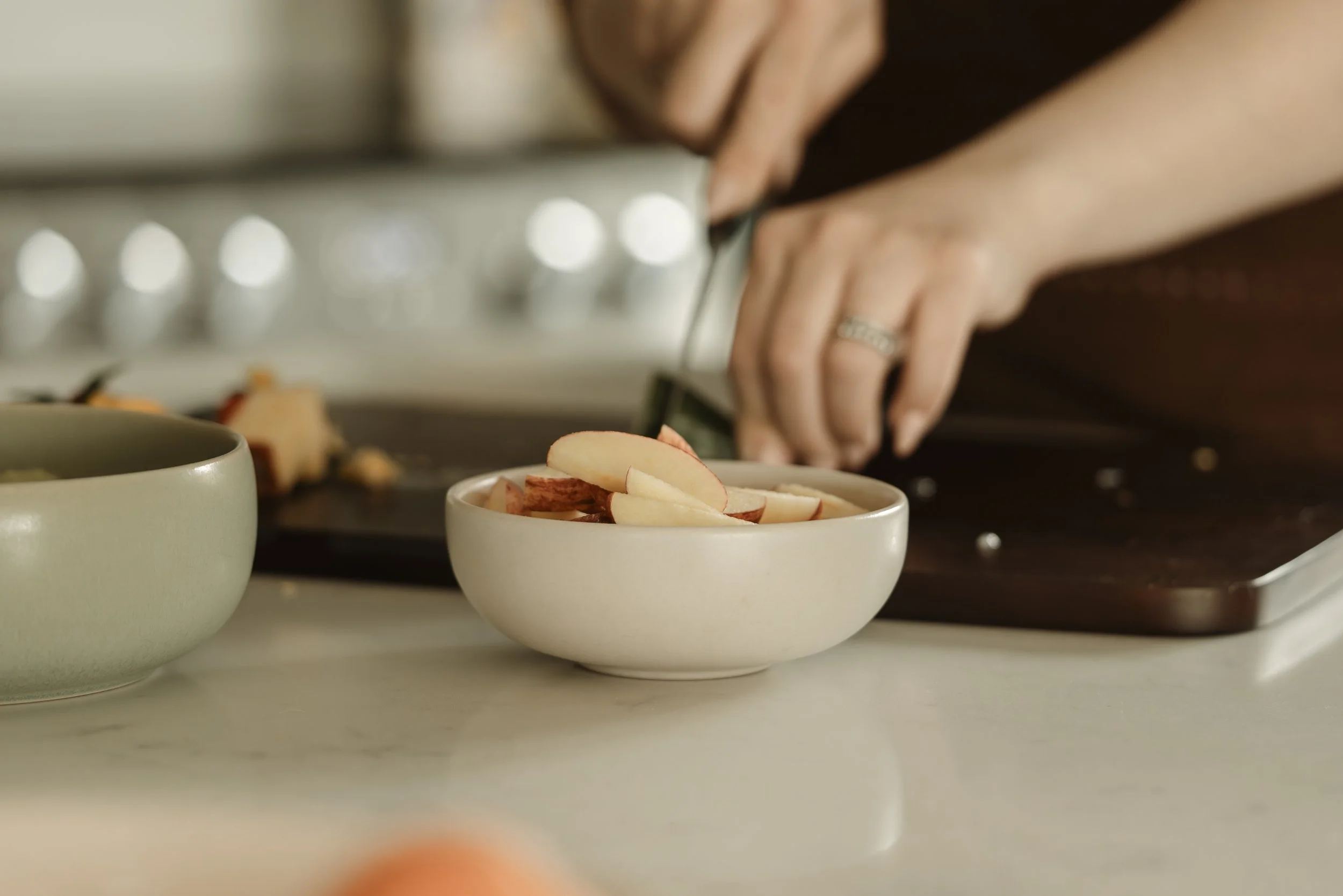 Person slicing apples on a cutting board in a kitchen with bowls of sliced apples in the foreground.