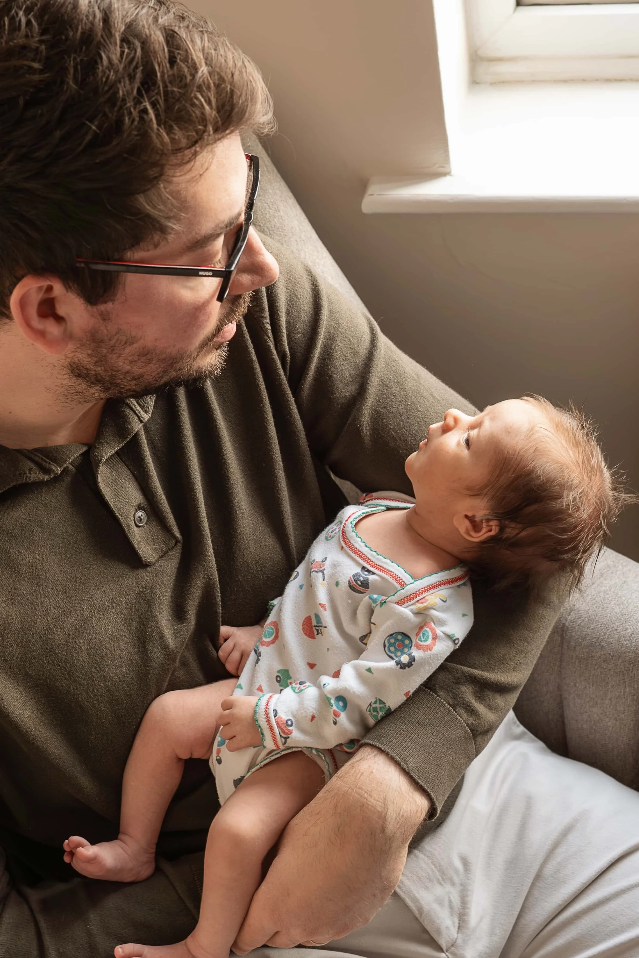 A man with glasses holding a baby girl while sitting on a couch near a window.