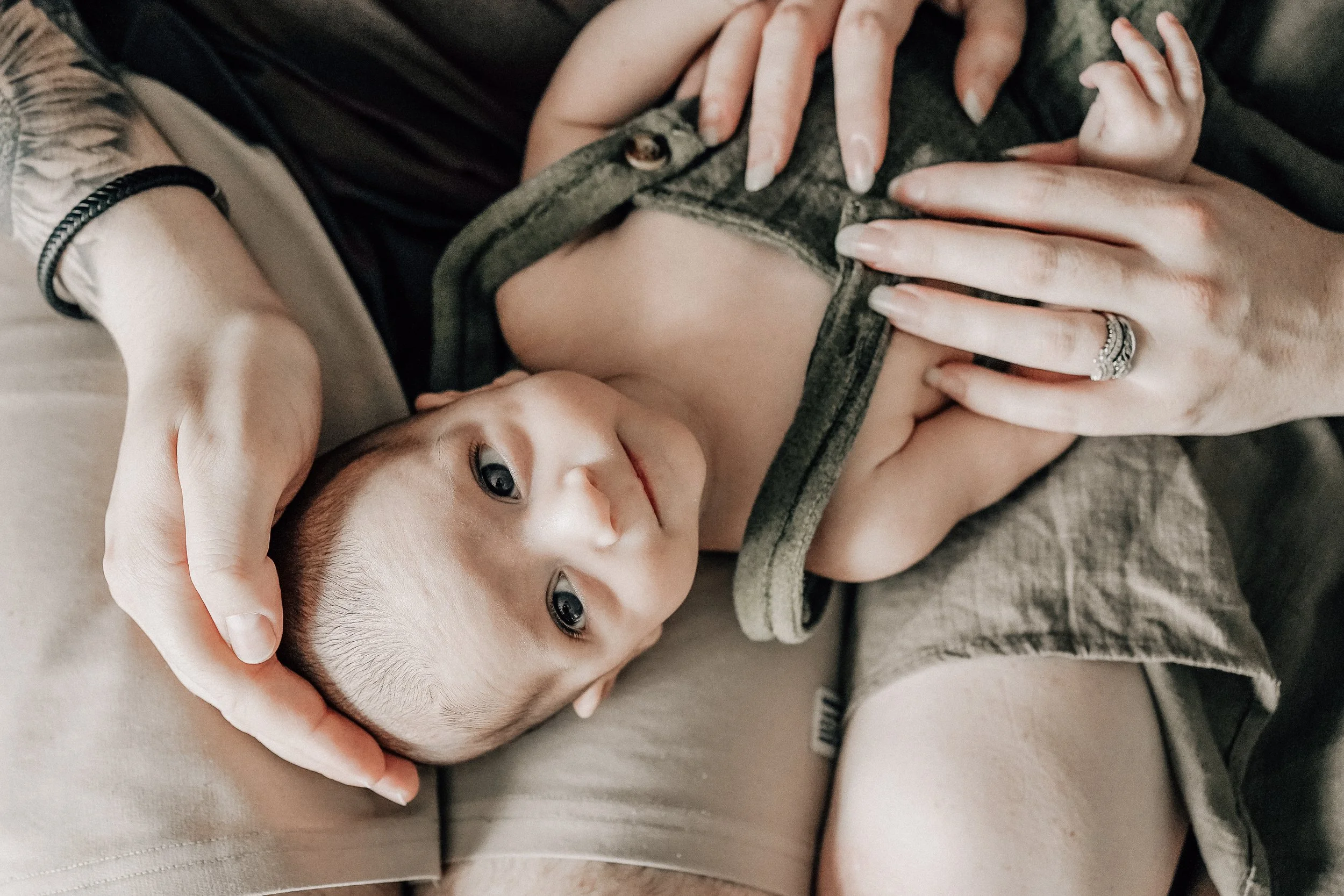 A baby lying on an adult's lap, with the adult gently holding the baby's head and chest. The baby is looking up with a slight smile, and the adult's hand is gently resting on the baby's chest while the other hand supports the baby's head.