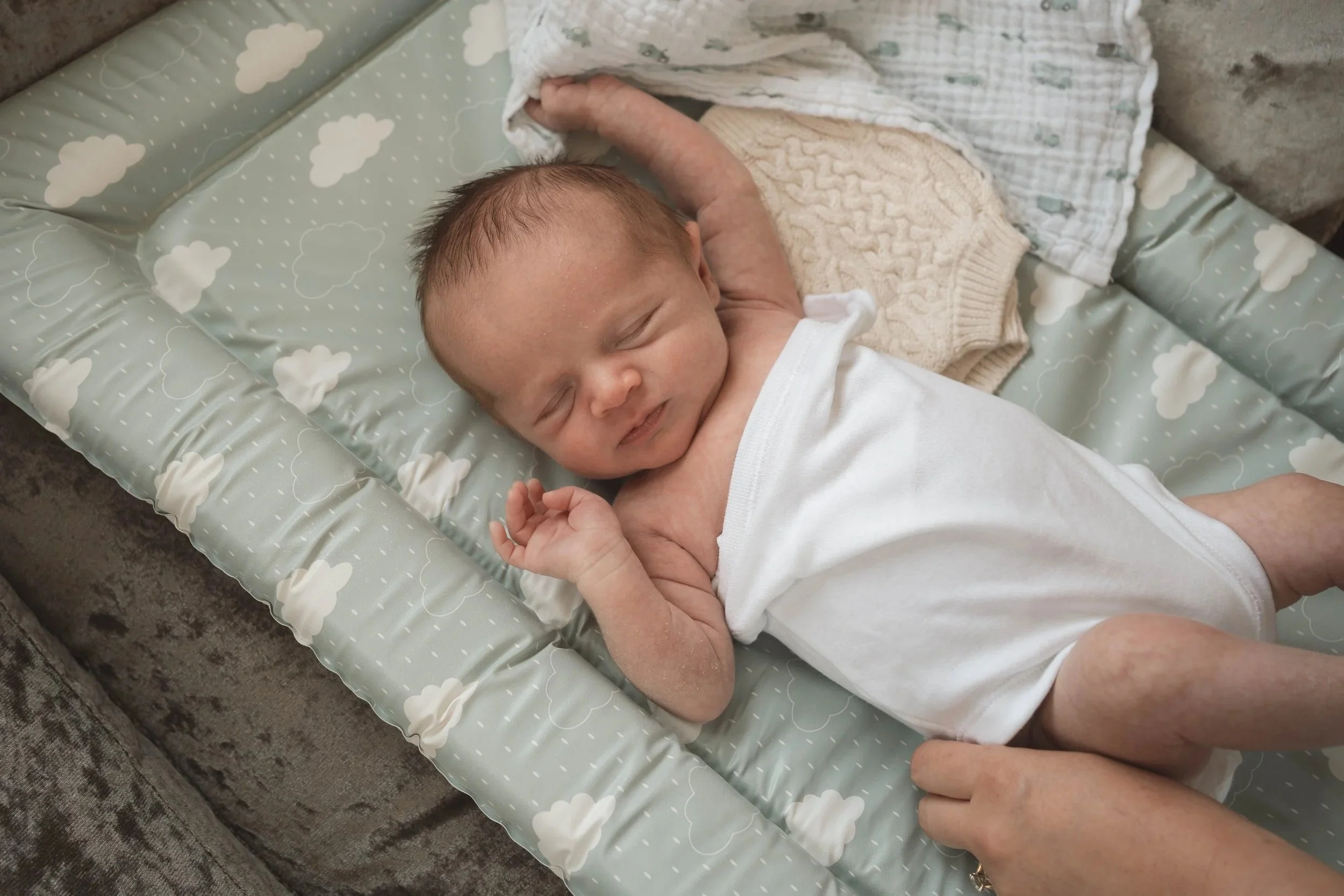 A sleeping baby lying on a cushioned mattress with a cloud pattern, wearing a white onesie, with a blanket nearby.
