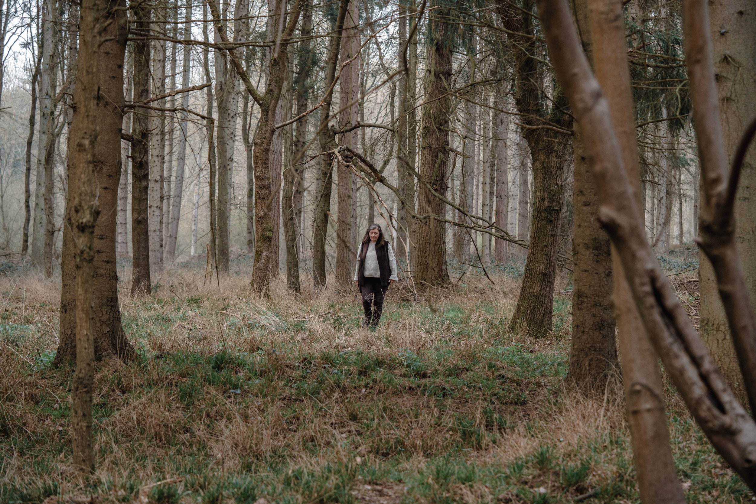 A woman walking through a dense forest with tall trees and dry grass on the ground.