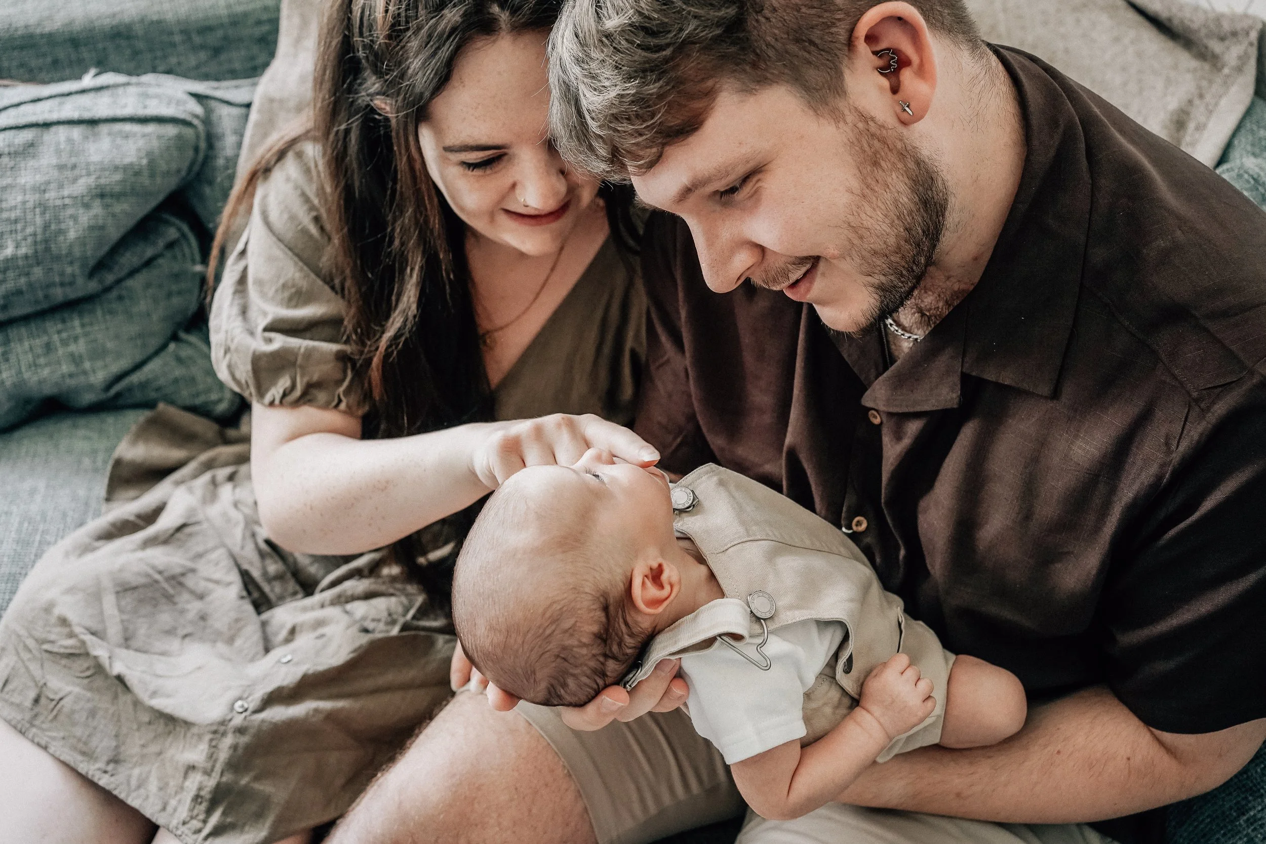 A couple sitting on a couch holding a baby, smiling and looking affectionately at the baby.