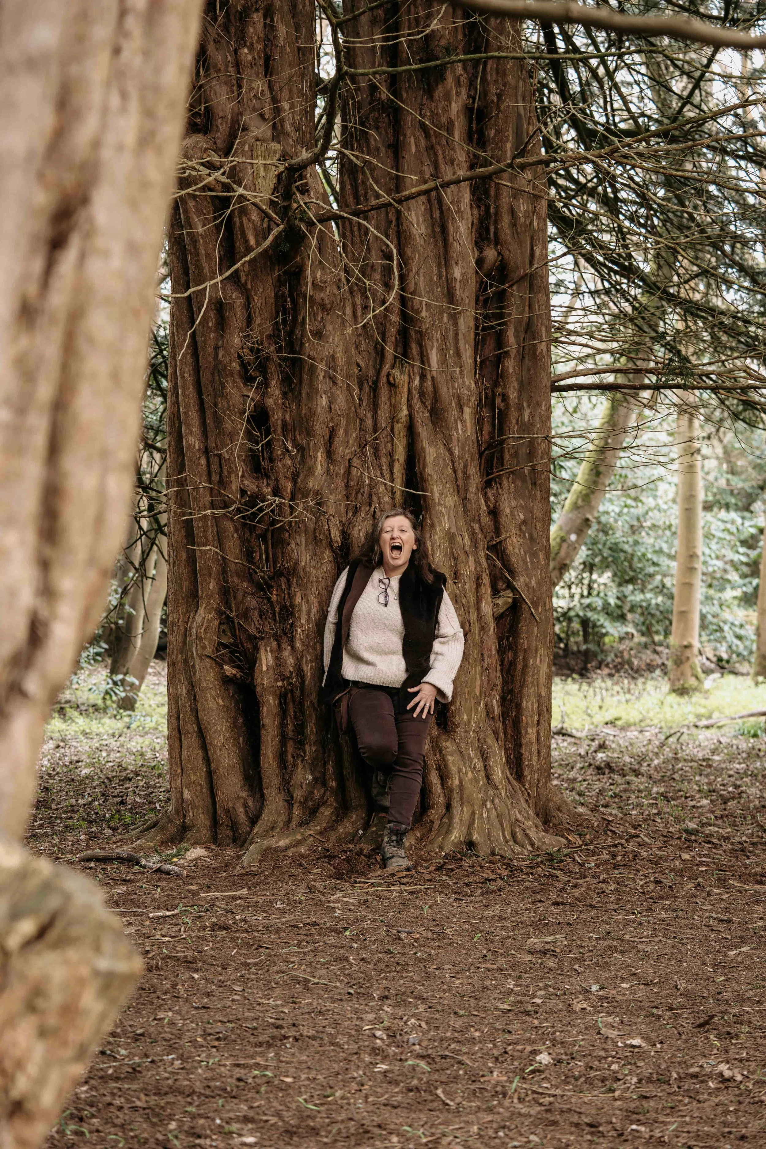 A woman with long dark hair, wearing a beige sweater, dark pants, and hiking boots, stands leaning against a large tree with reddish-brown bark in a wooded area. She appears to be laughing or smiling with her mouth open and has glasses hanging from h