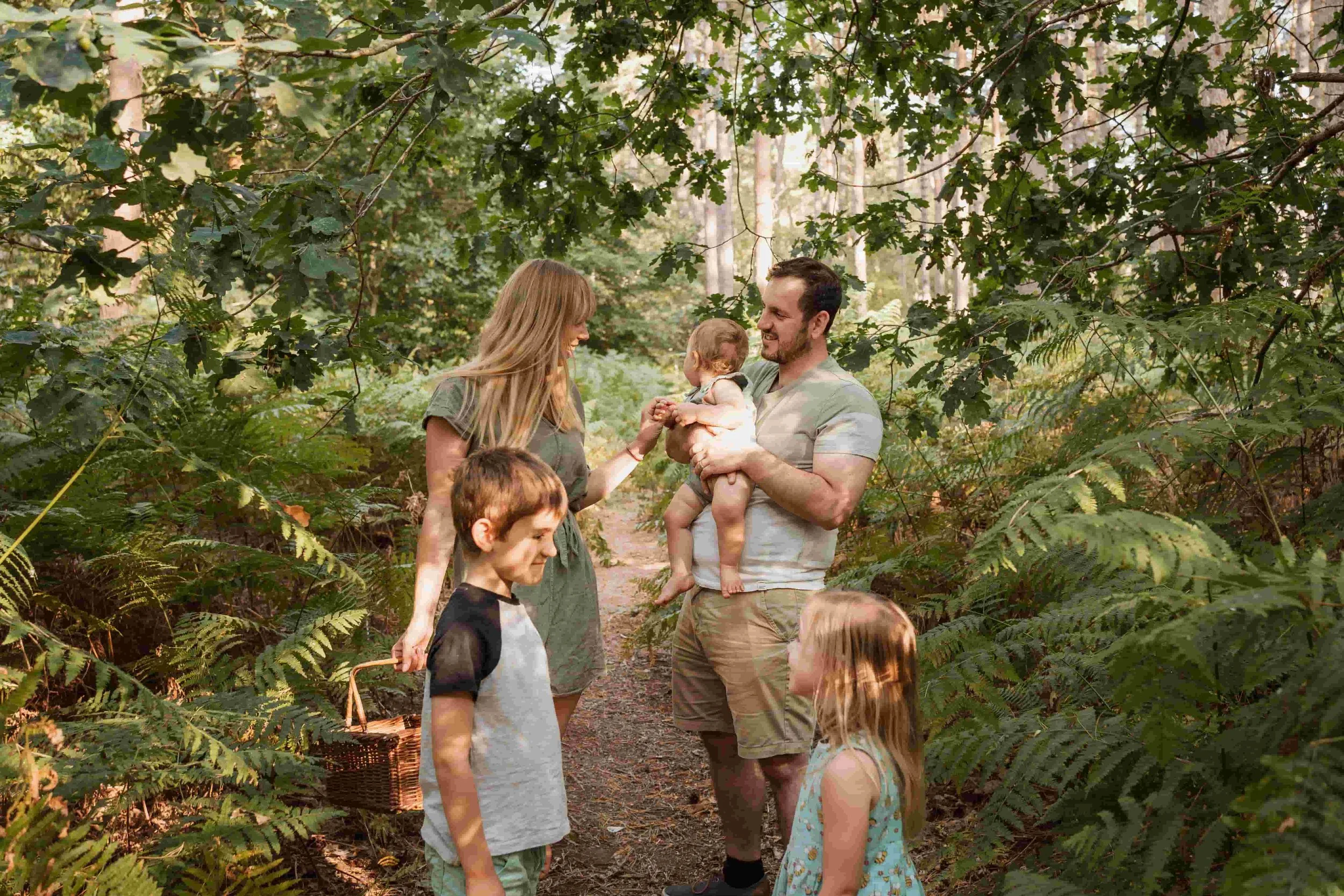 A family of five, including two adults and three children, enjoying a walk in a wooded area during daytime. The adults are smiling at each other, and the man is holding a young child.