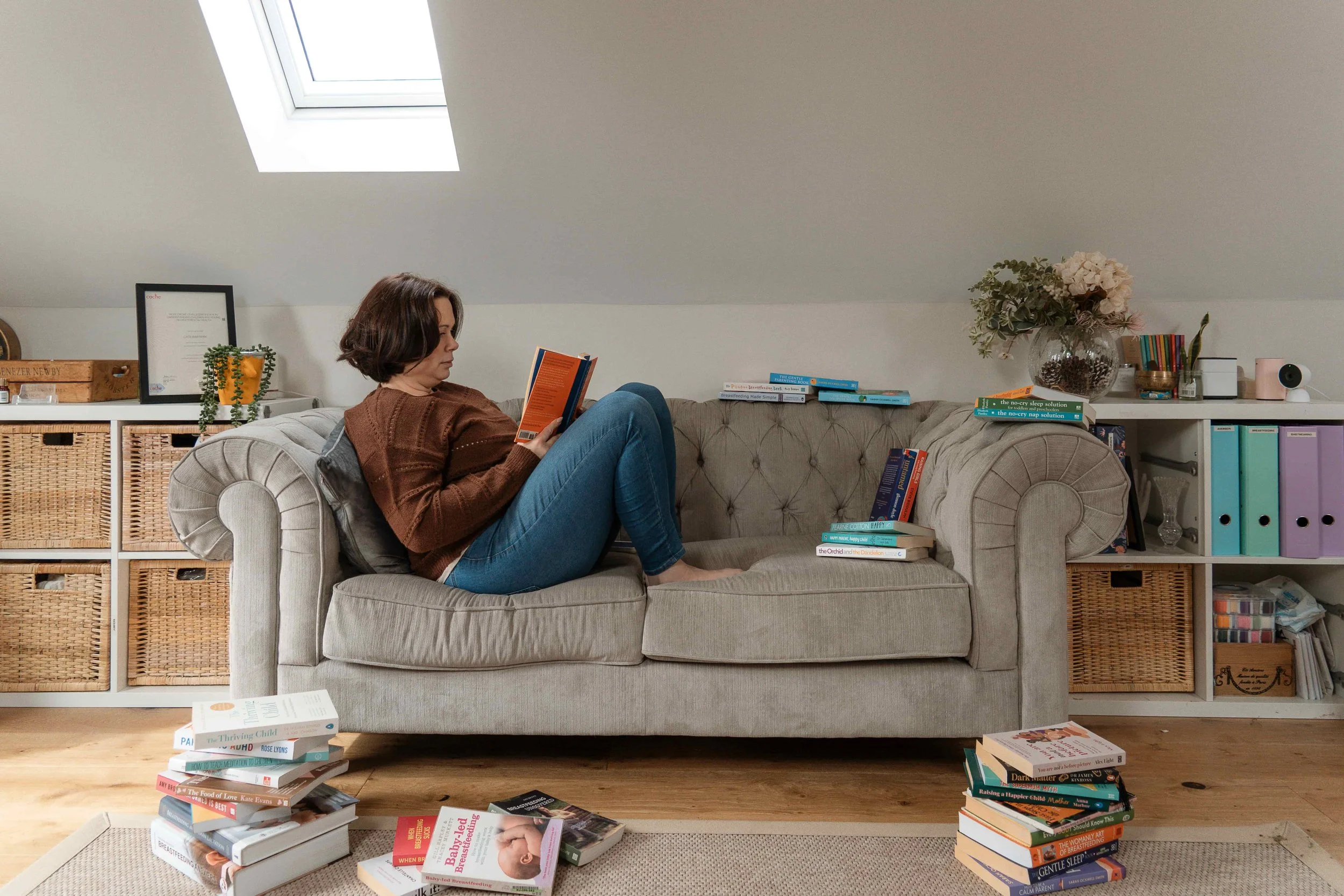 A woman with short dark hair reading a book while sitting on a beige sofa in a room with a white ceiling and a large skylight window. The room has shelves filled with books, binders, and decorative items, and a potted plant on one of the shelves. Sta