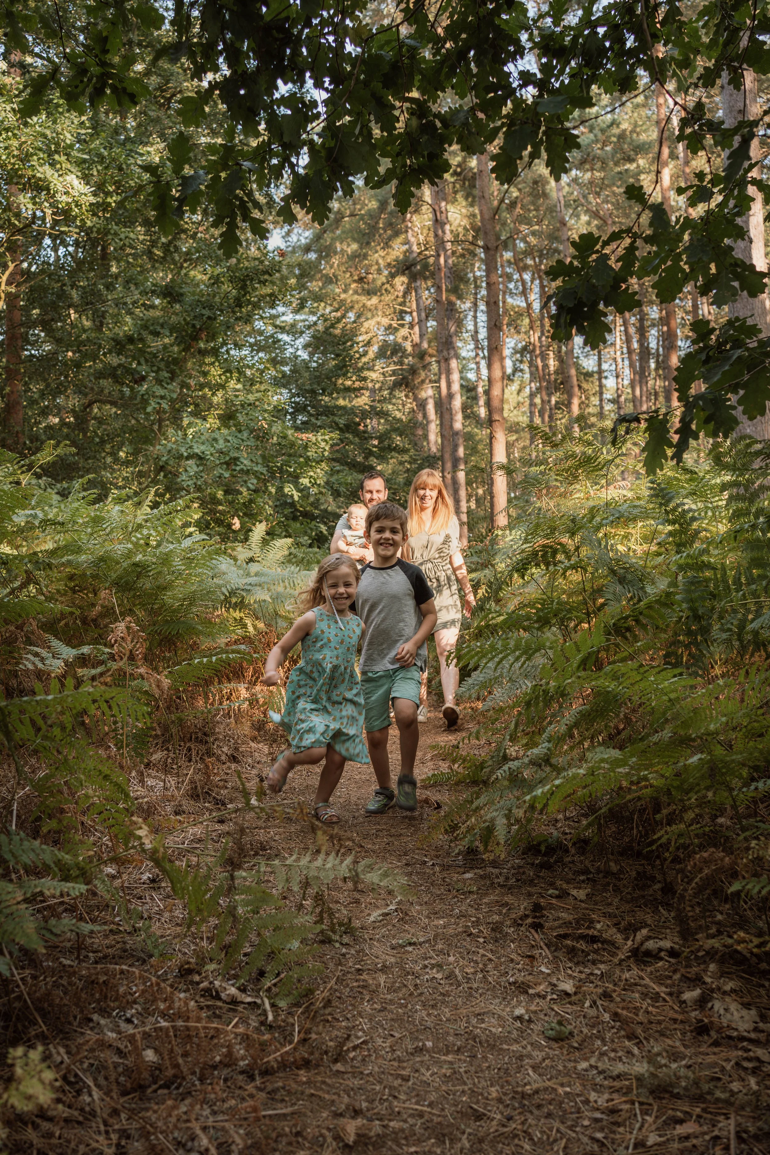 A family of five, including two children, walking and playing on a dirt trail through a wooded forest with tall trees and green ferns.