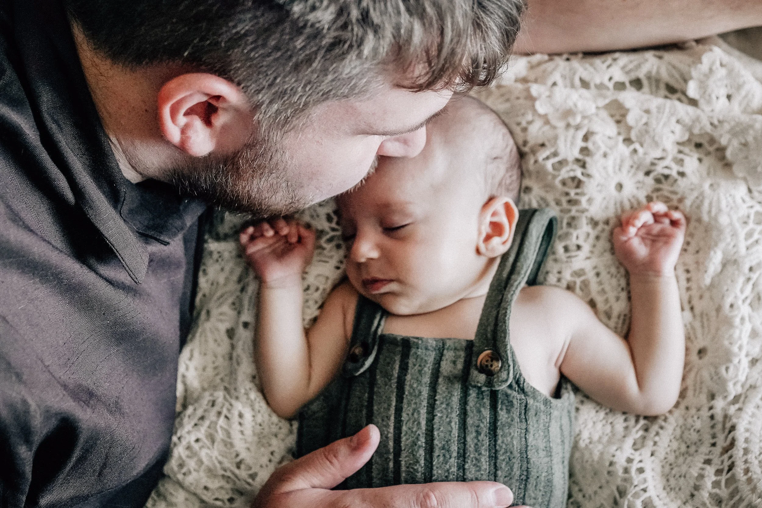 A man and a baby lying face to face on a lace blanket, with the man gently touching the baby's chest.