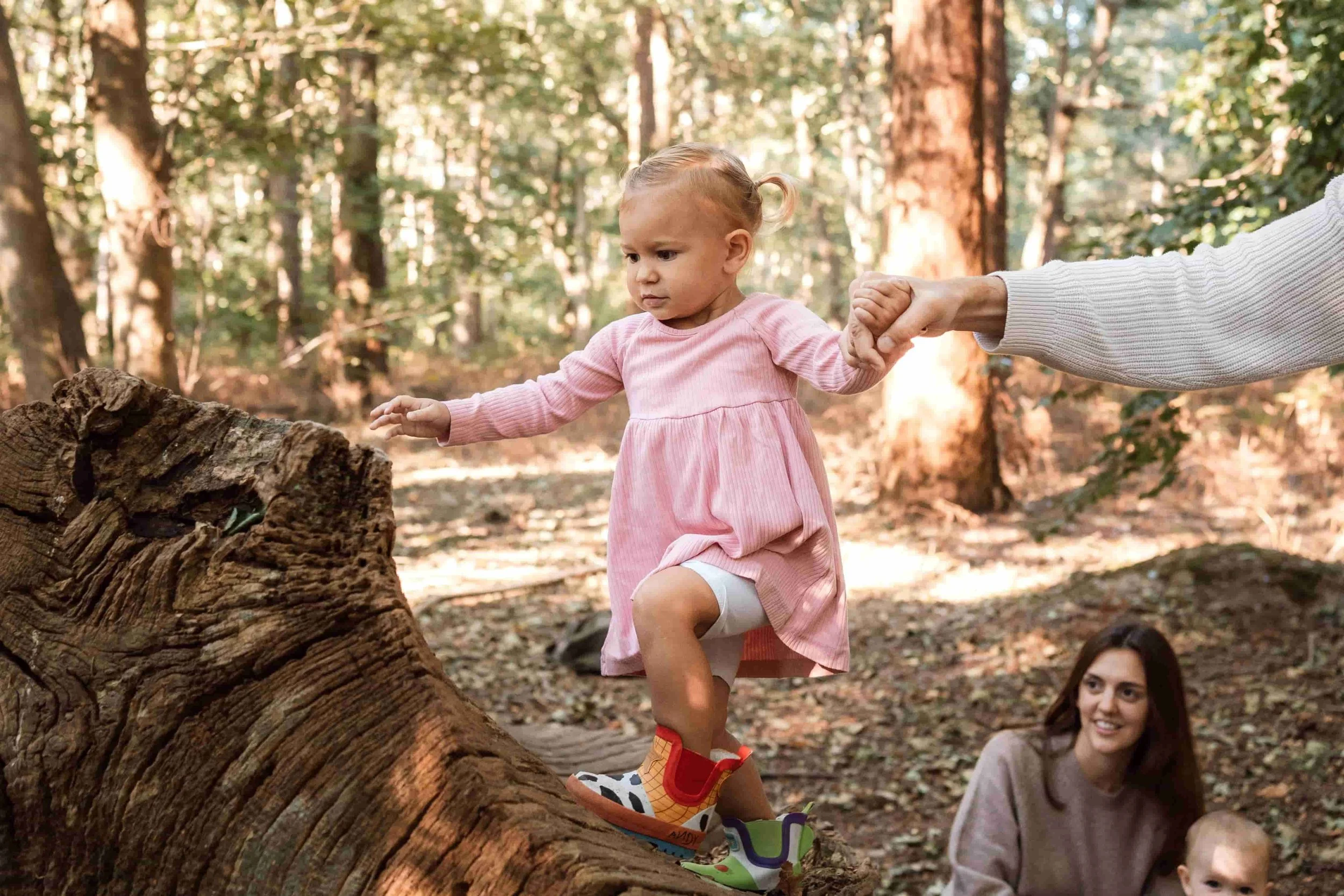 A young girl in a pink dress helps hold the hand of an adult as she balances on a large fallen tree in a forest. The girl wears colorful sneakers and has her hair styled in a small ponytail. An adult woman with long brown hair and a beige sweater is 