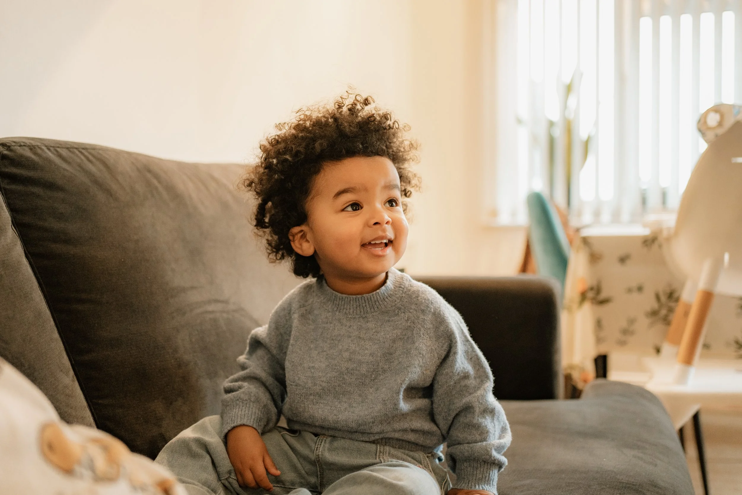 A young child with curly hair sitting on a gray couch, smiling and looking to the side in a well-lit room.