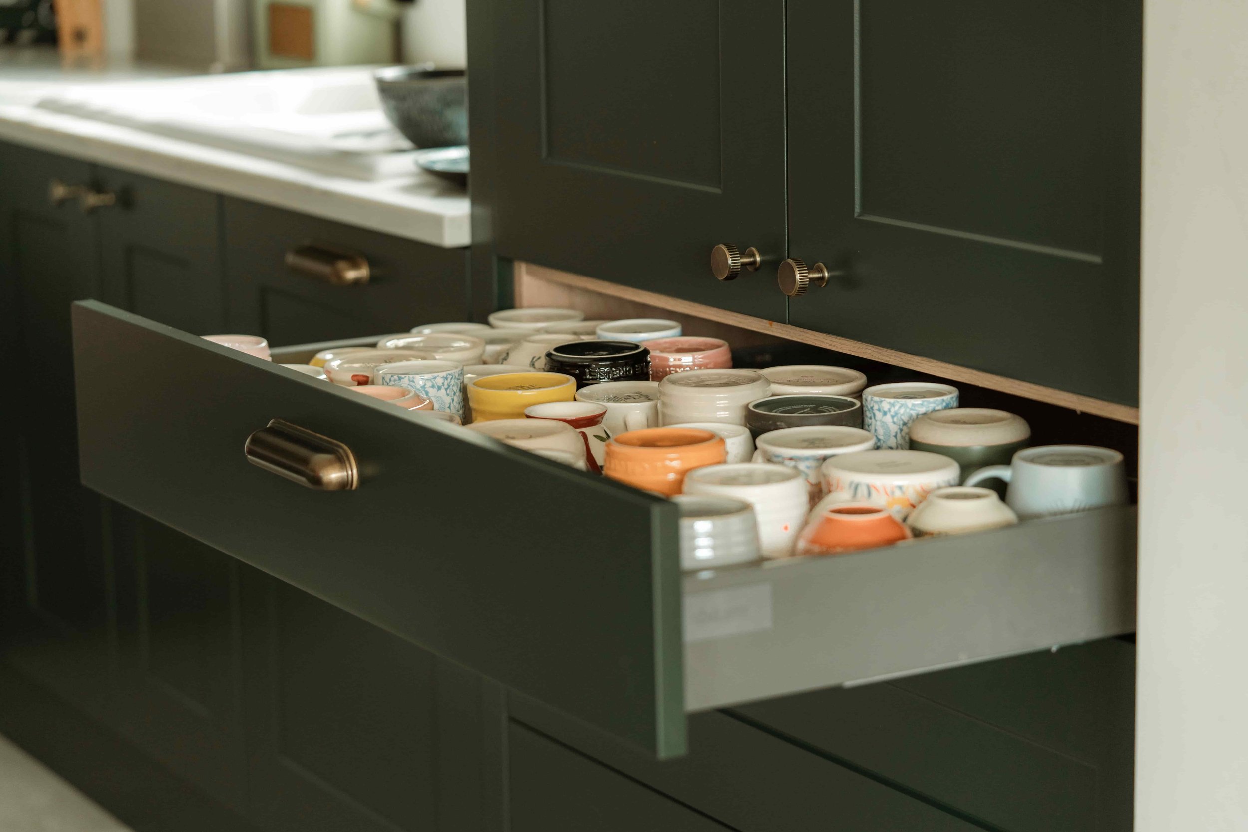 Open drawer filled with various colorful ceramic cups and mugs. Dark green cabinet and countertop with a bowl in the background.