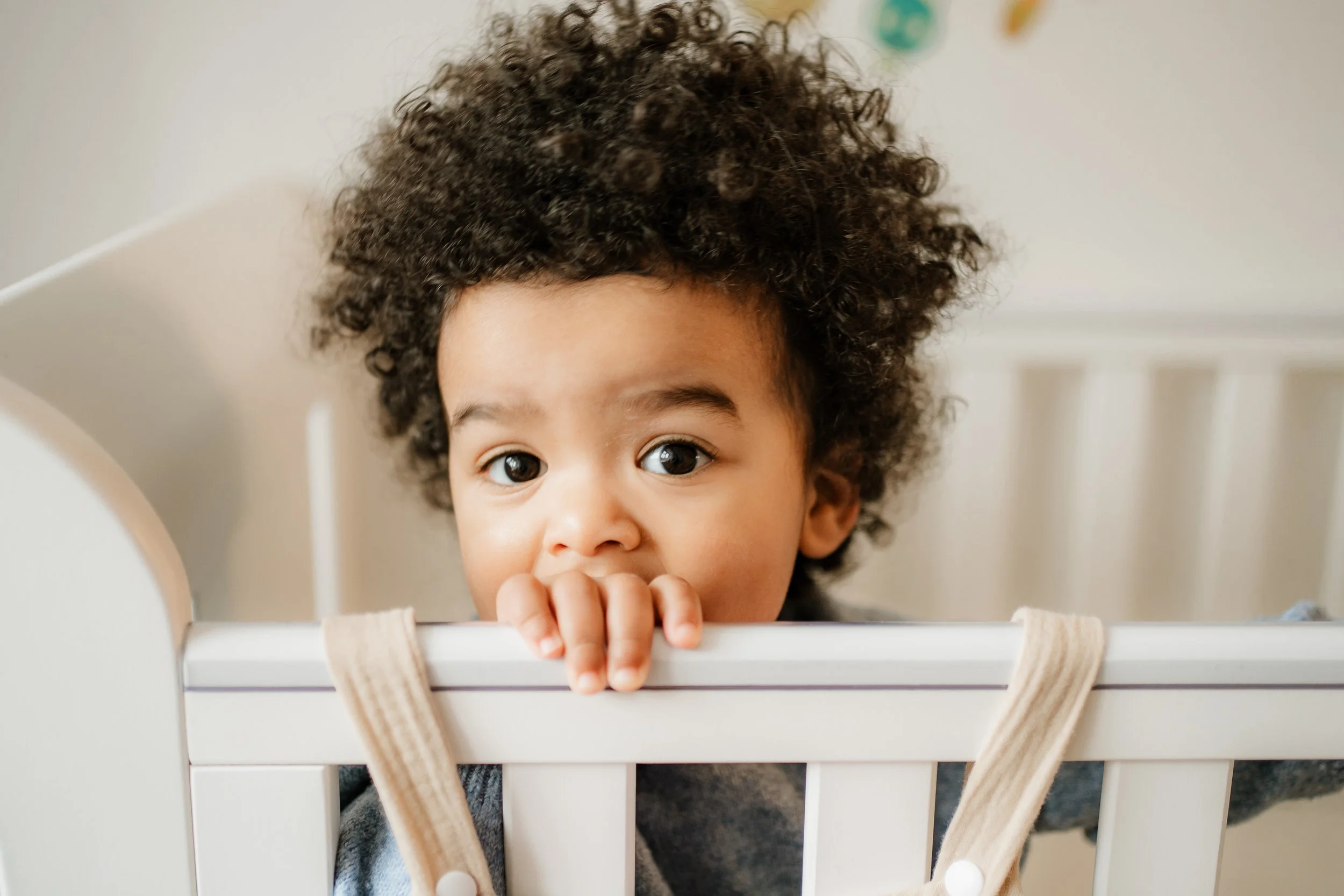 A young child with curly hair peeking over the side of a white crib, holding onto the top rail with one hand, looking at the camera with wide eyes.