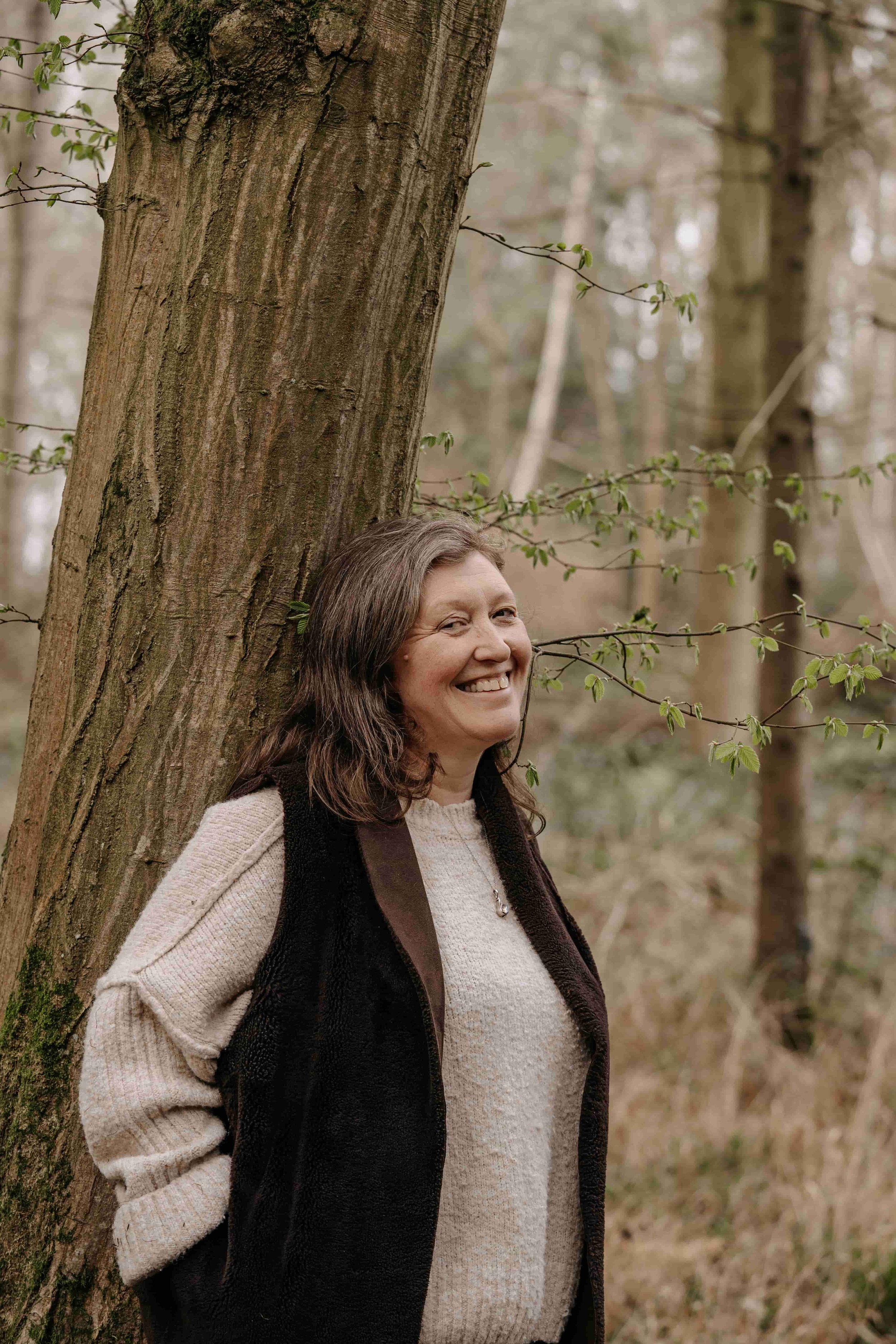 A woman with long brown hair smiling outdoors, leaning against a tree trunk with green leaves and trees in the background.