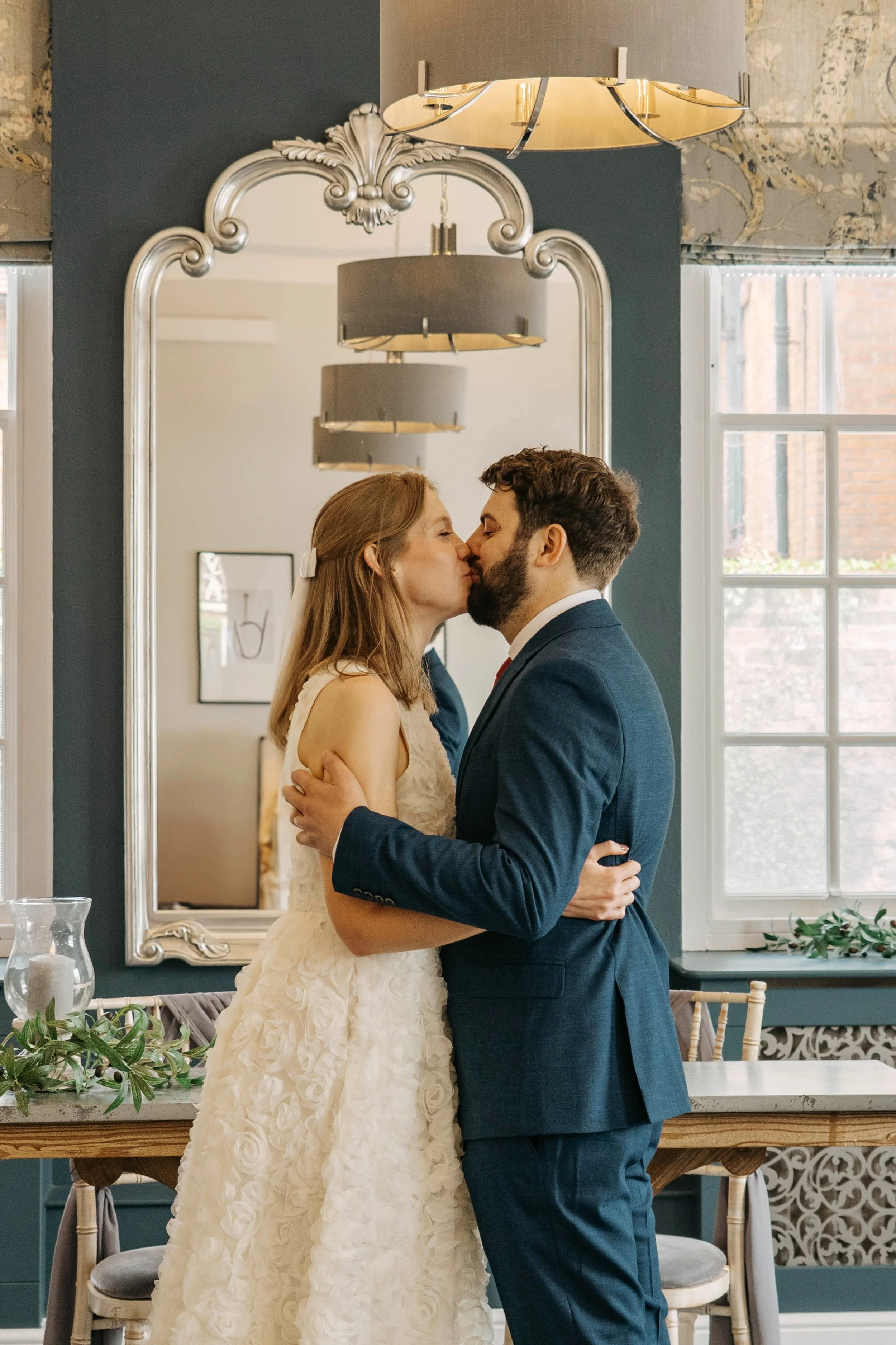 A couple in wedding attire kissing in front of a mirror inside a decorated room.