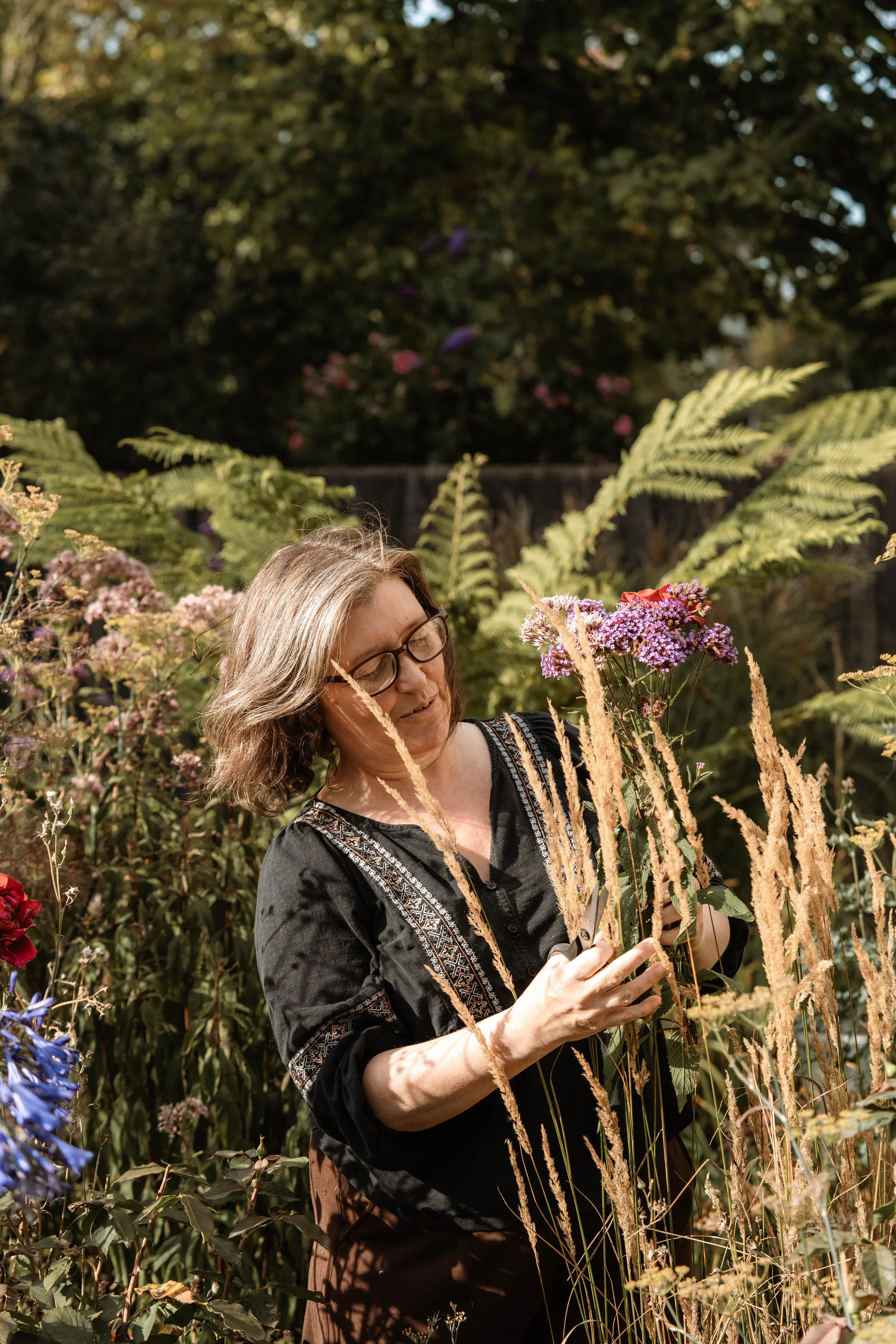 A woman with glasses and short, wavy hair, dressed in a black embroidered top, trims pink and purple flowers in a garden with tall grasses and various plants.