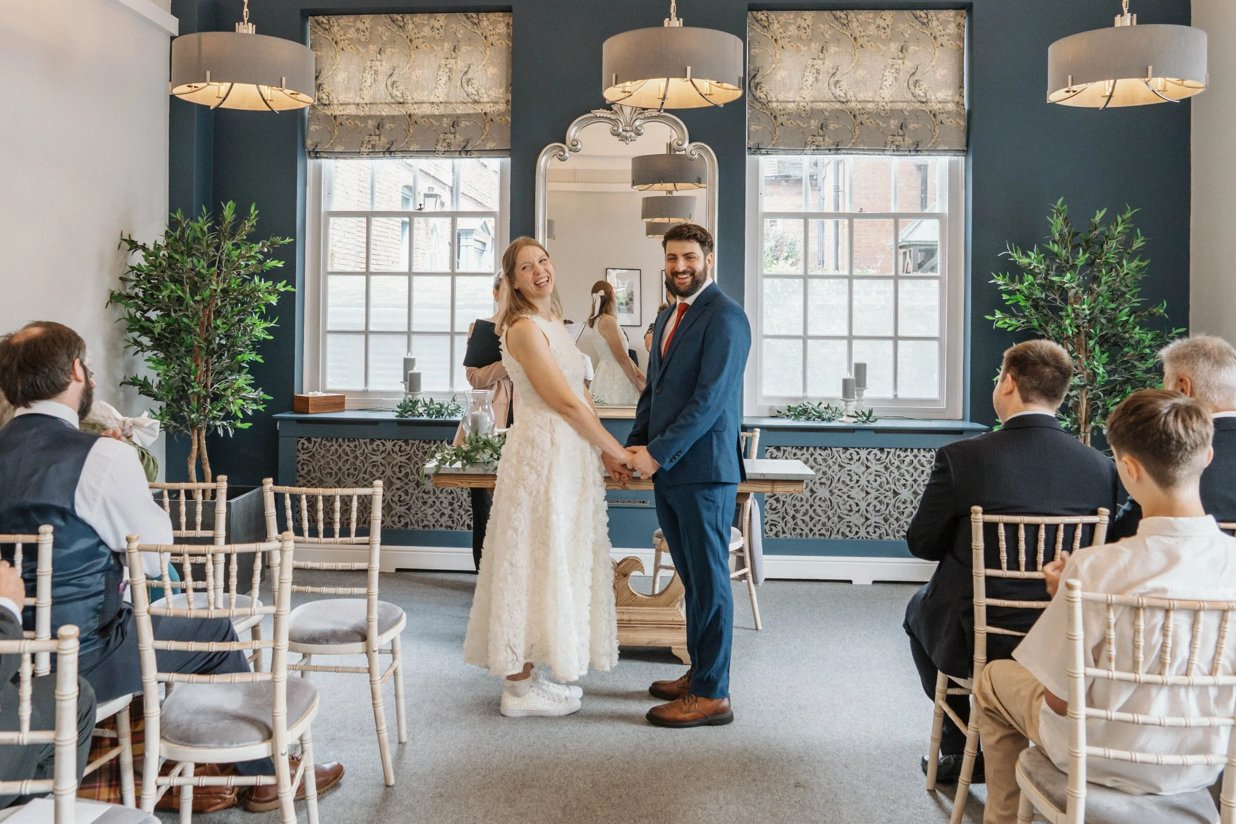 A wedding ceremony with a bride and groom holding hands and smiling at each other, surrounded by seated guests in a decorated indoor venue with large windows, plants, and modern lighting fixtures.