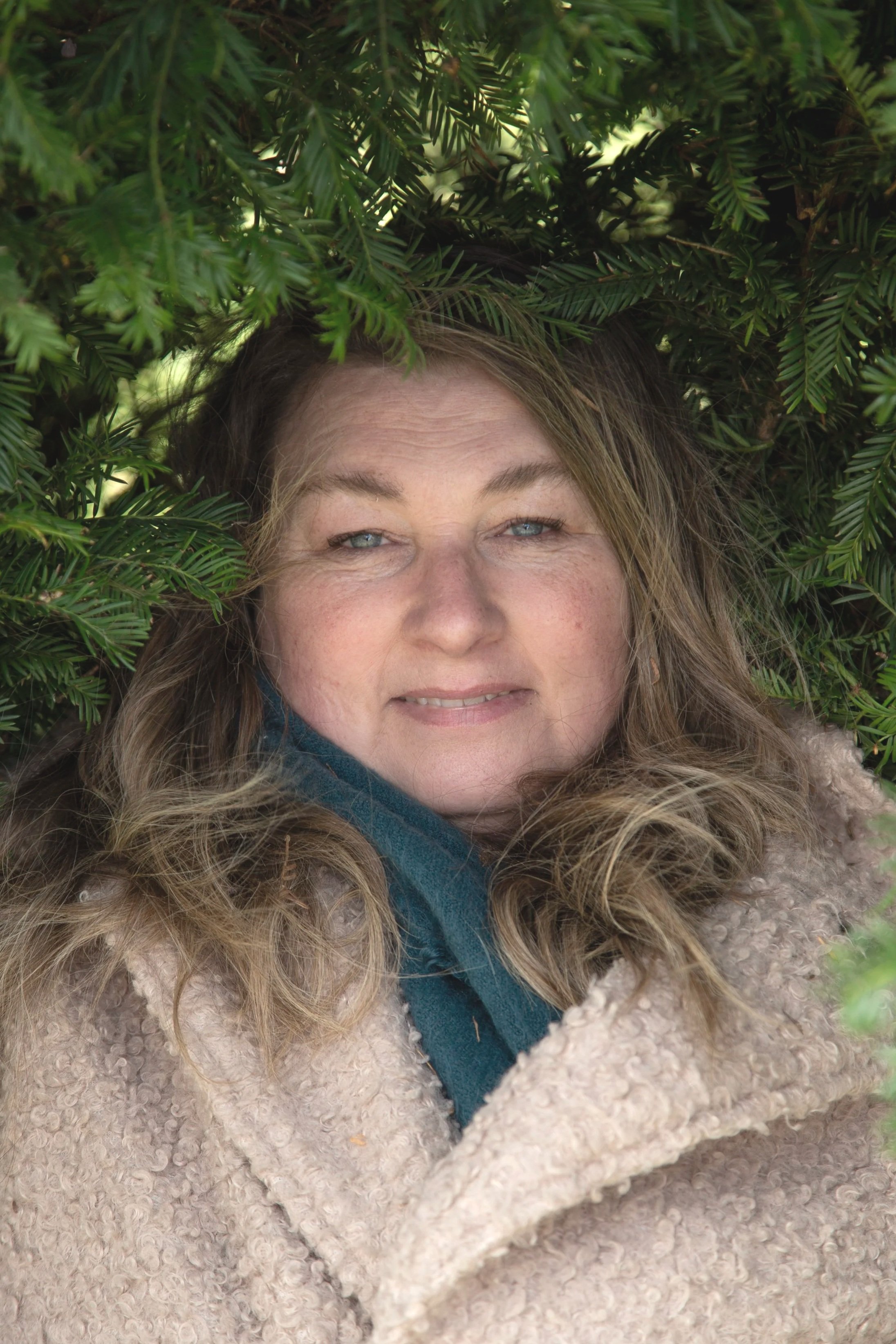 A woman with wavy blonde hair and blue eyes, wearing a beige fleece coat and teal scarf, lying amongst green leafy branches.