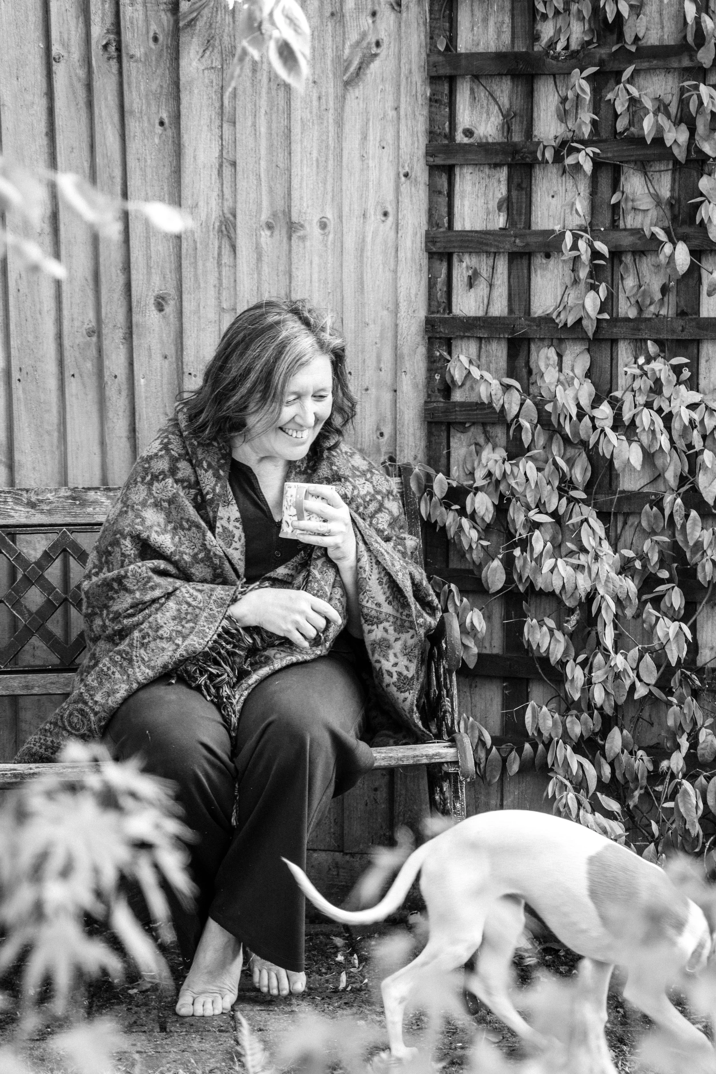 A woman sitting on a wooden bench outdoors, smiling and holding a mug, with a dog in front of her, surrounded by a wooden fence and foliage.