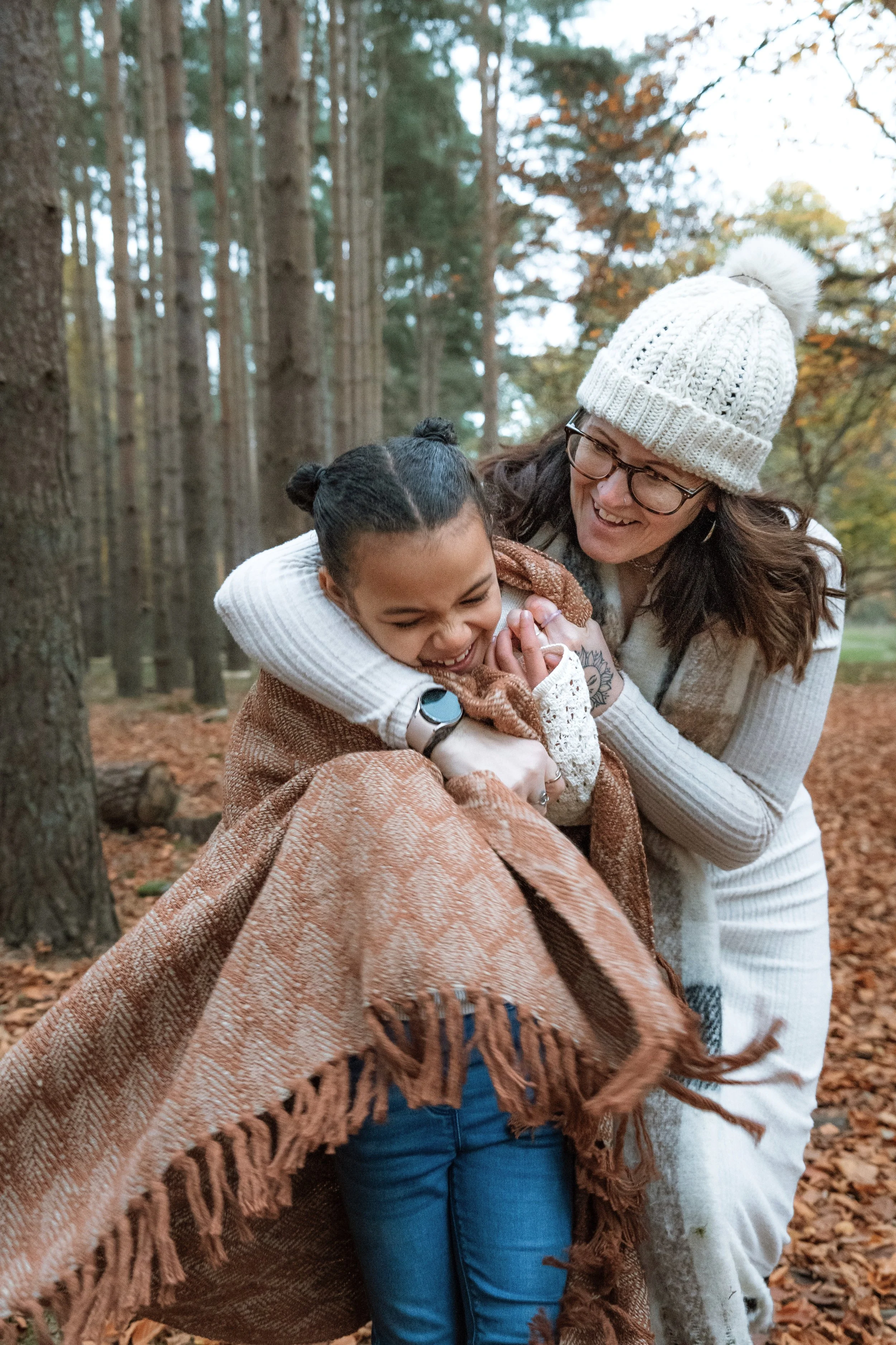 A woman and a girl walking together in a forest with autumn leaves on the ground, both dressed warmly for fall.