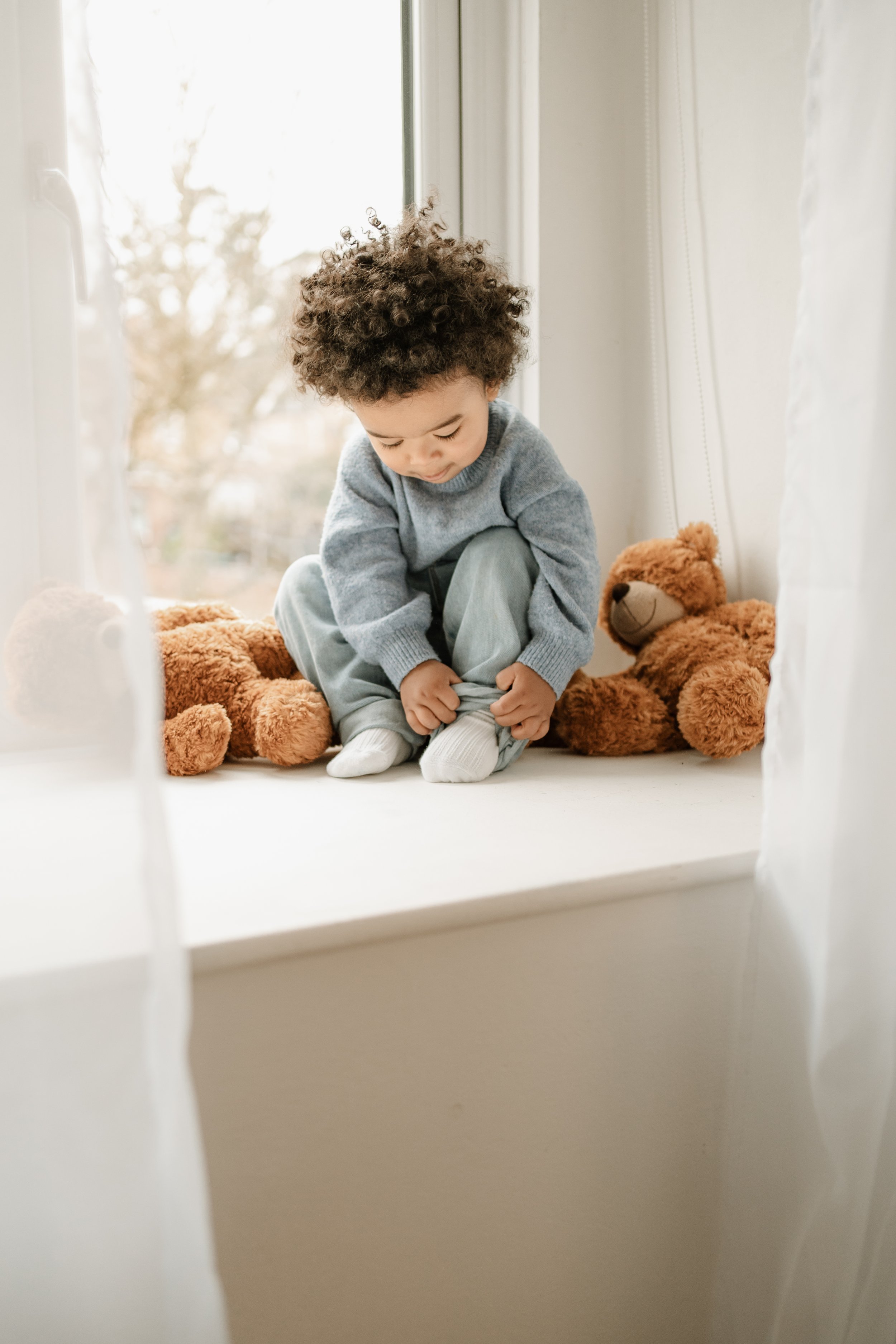 A young child with curly hair sitting on a windowsill, tying the laces of his white shoes, surrounded by stuffed teddy bears.