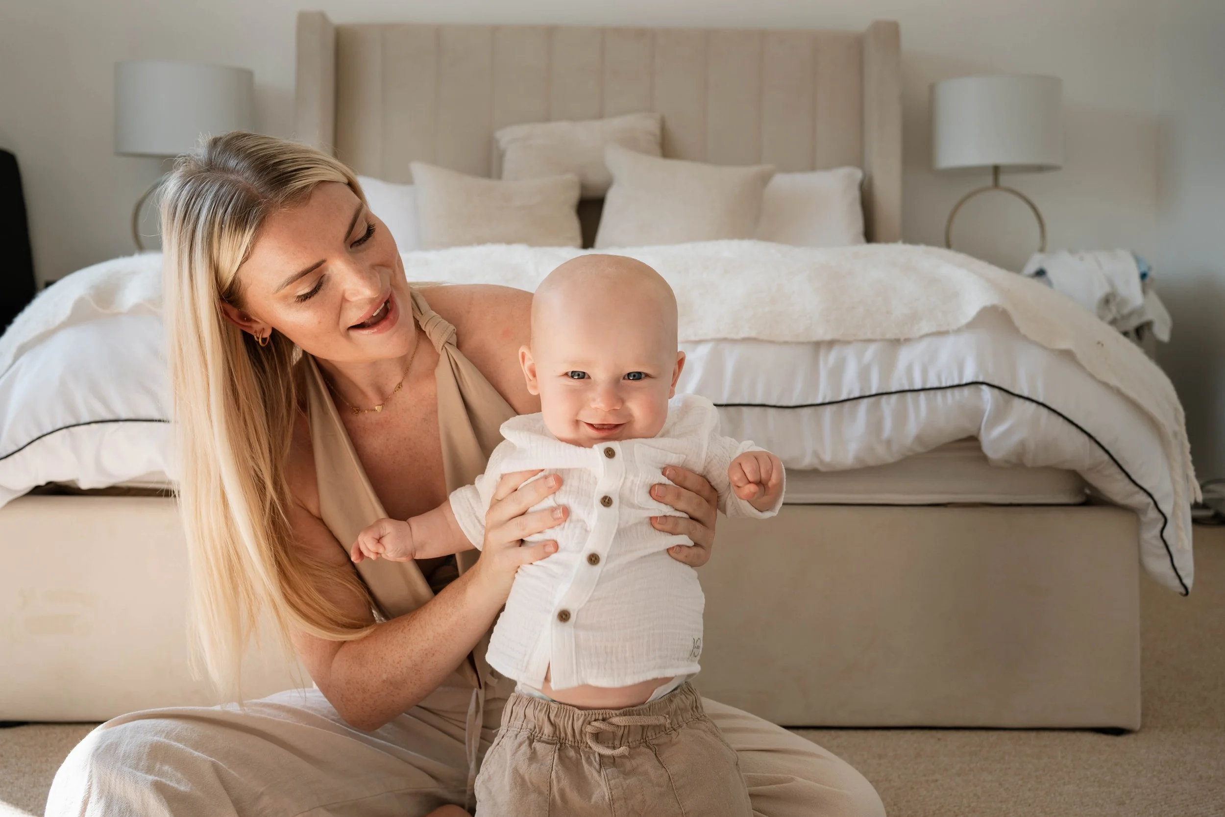 A woman with long blonde hair and a young boy with light skin and bald head smiling and playing together on a bed in a bedroom with neutral-colored decor.
