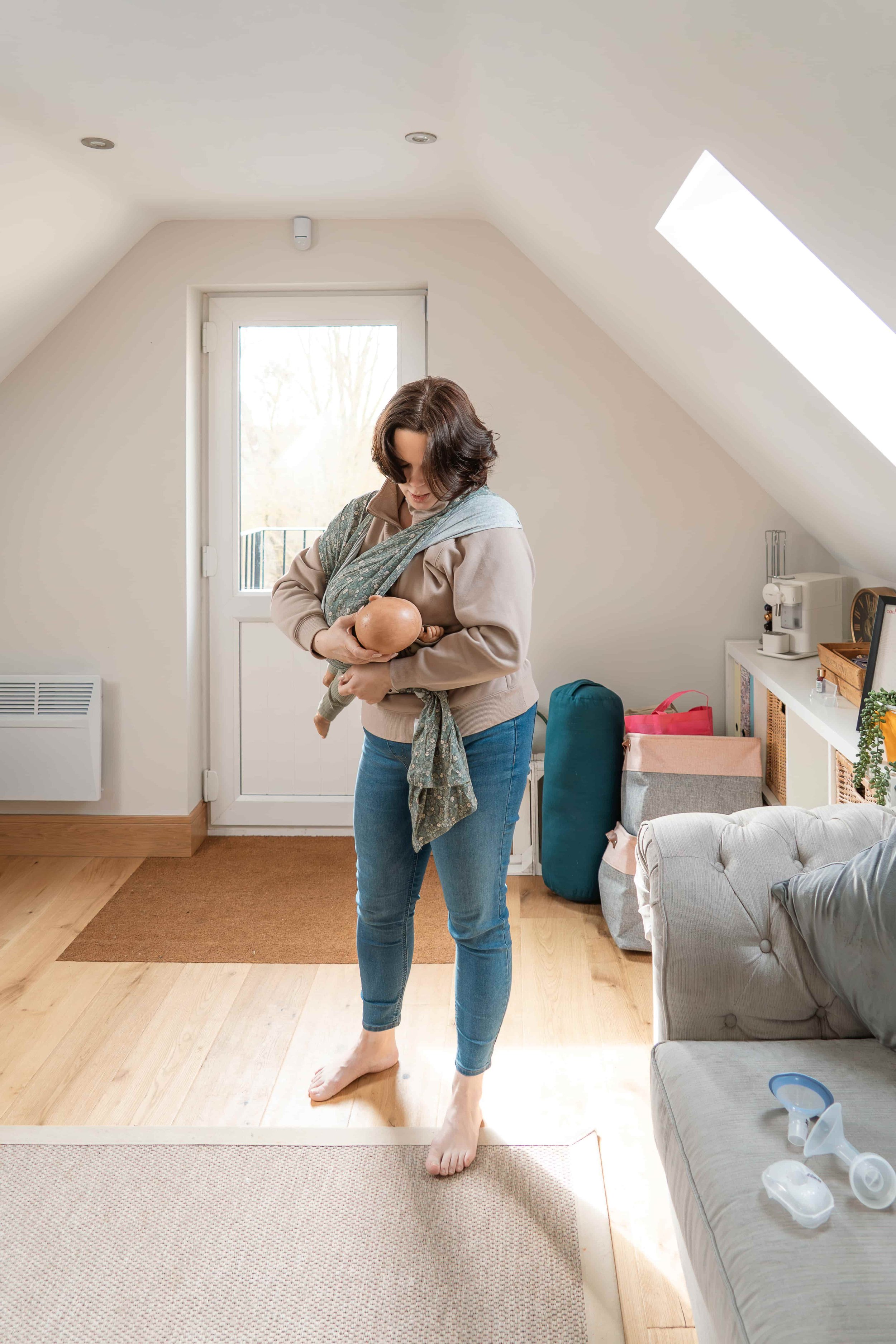 A woman standing barefoot on a beige rug in a bright room holding a baby, with a window and a slanted skylight above. The room has light-colored walls, wooden flooring, and furniture including a gray sofa, a white shelf, and some boxes and bags. Ther