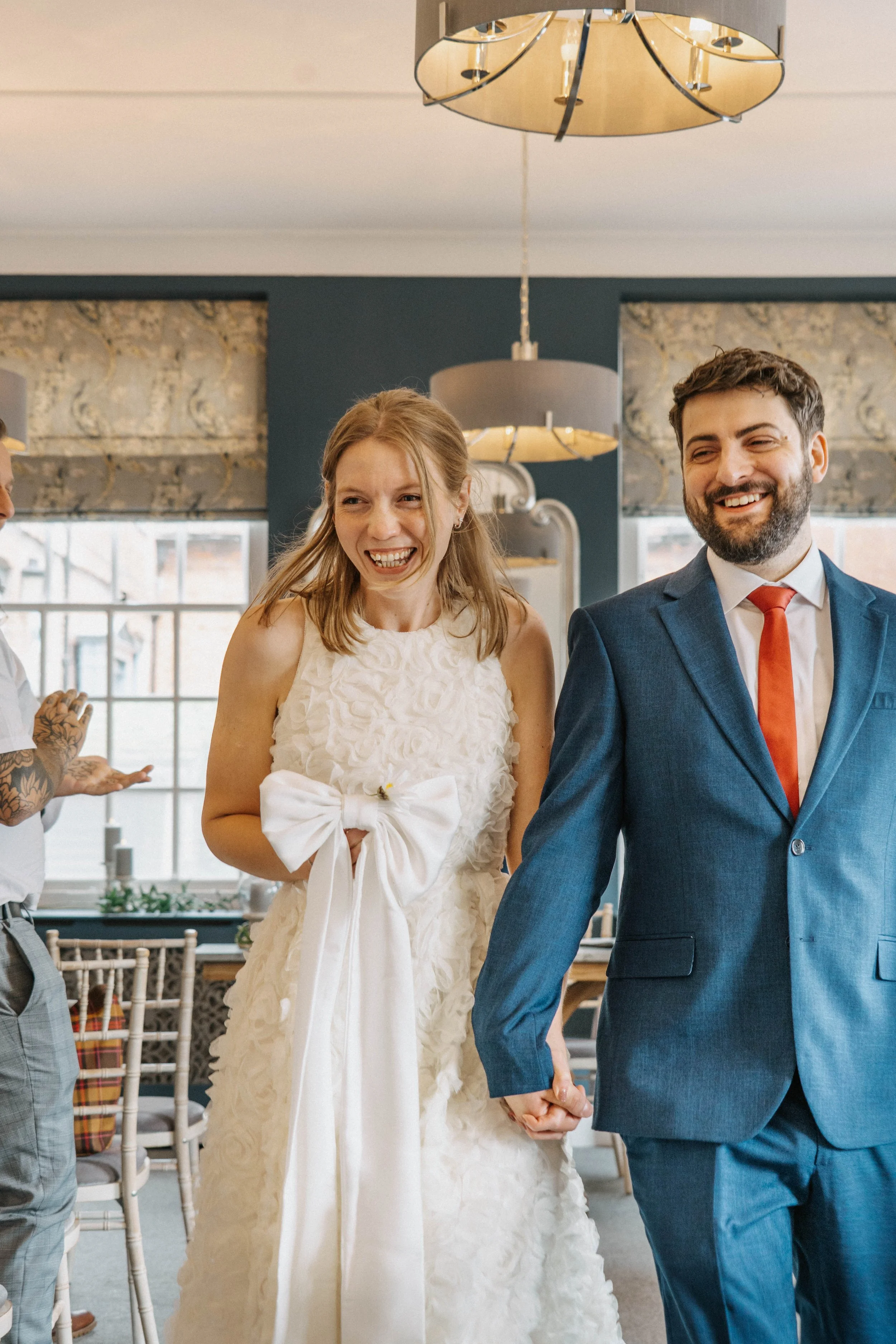 A happy couple at their wedding, the woman in a white dress and the man in a blue suit, holding hands and smiling, indoors with large windows and decorative lighting.