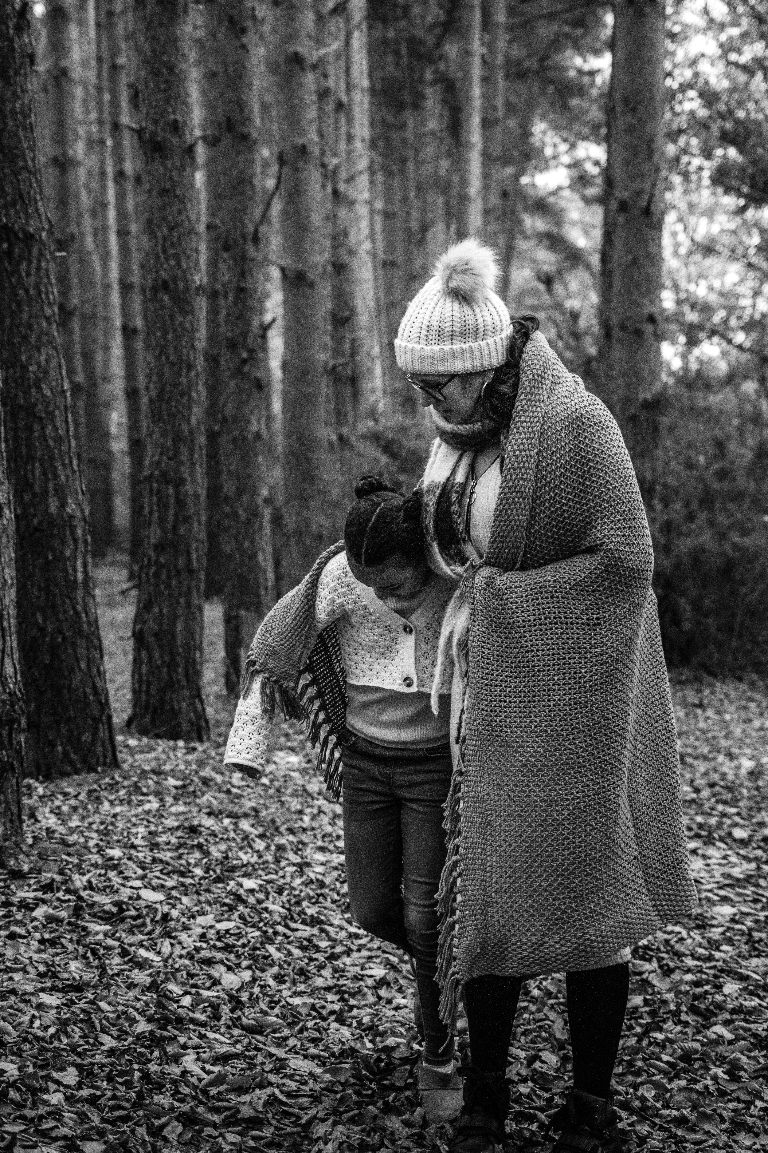 A woman and a girl walking through a forest on a leaf-covered path, both bundled in warm clothing and wrapped in a blanket.