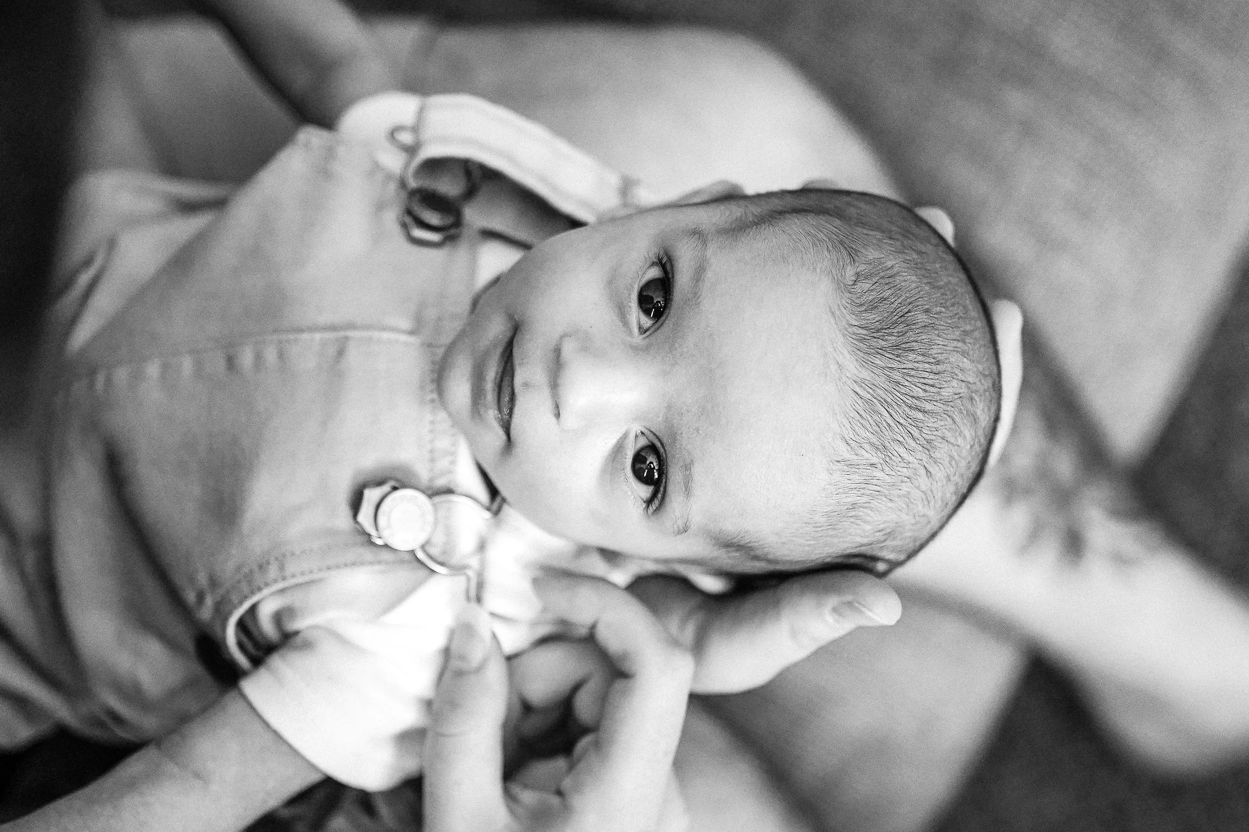 A young child with a slight smile, resting on an adult's hand, looking upwards, in black and white.