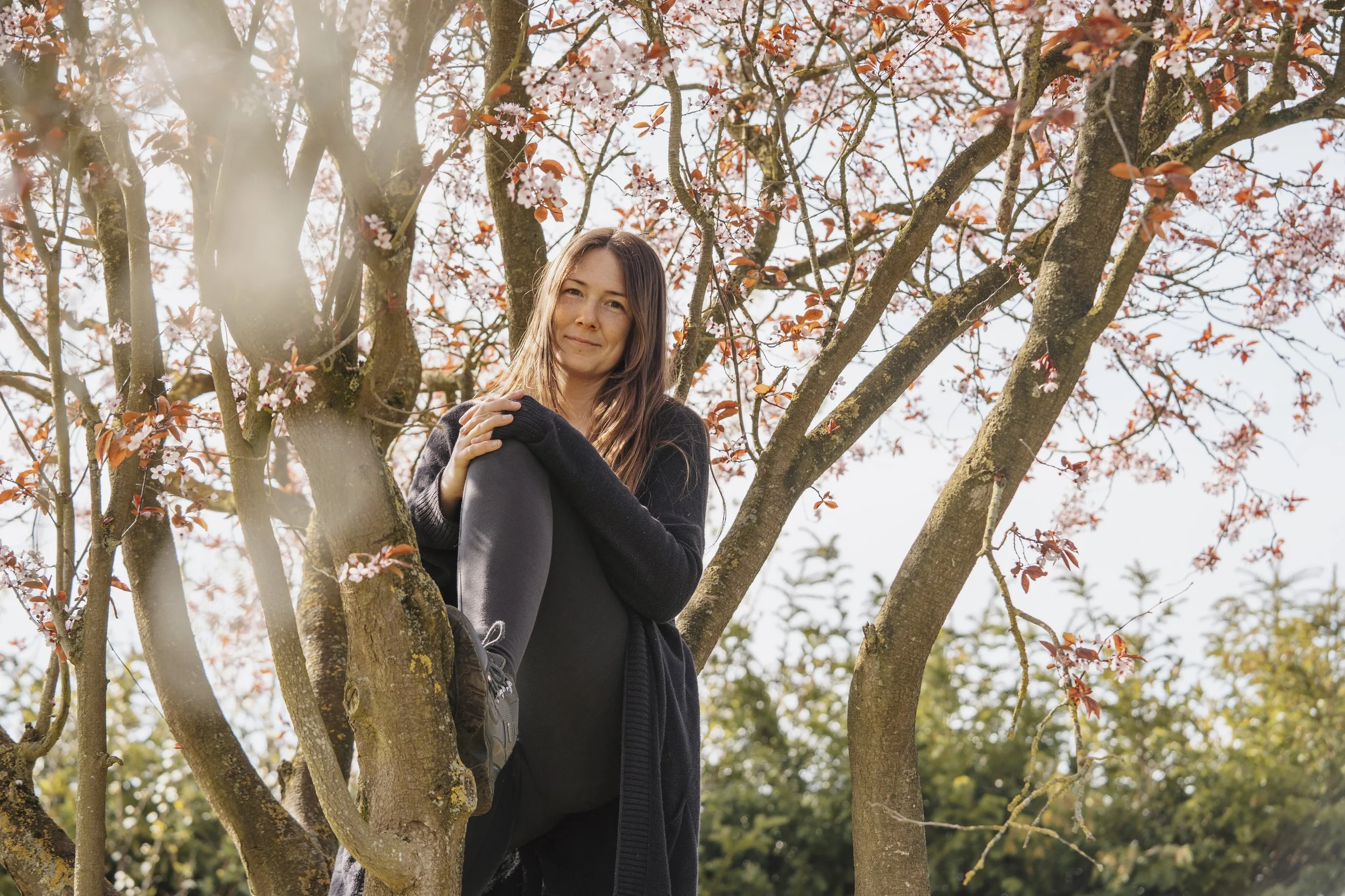 A woman in black sitting on a tree branch among pink and white blossoms, smiling and looking at the camera with sunlight filtering through the branches.
