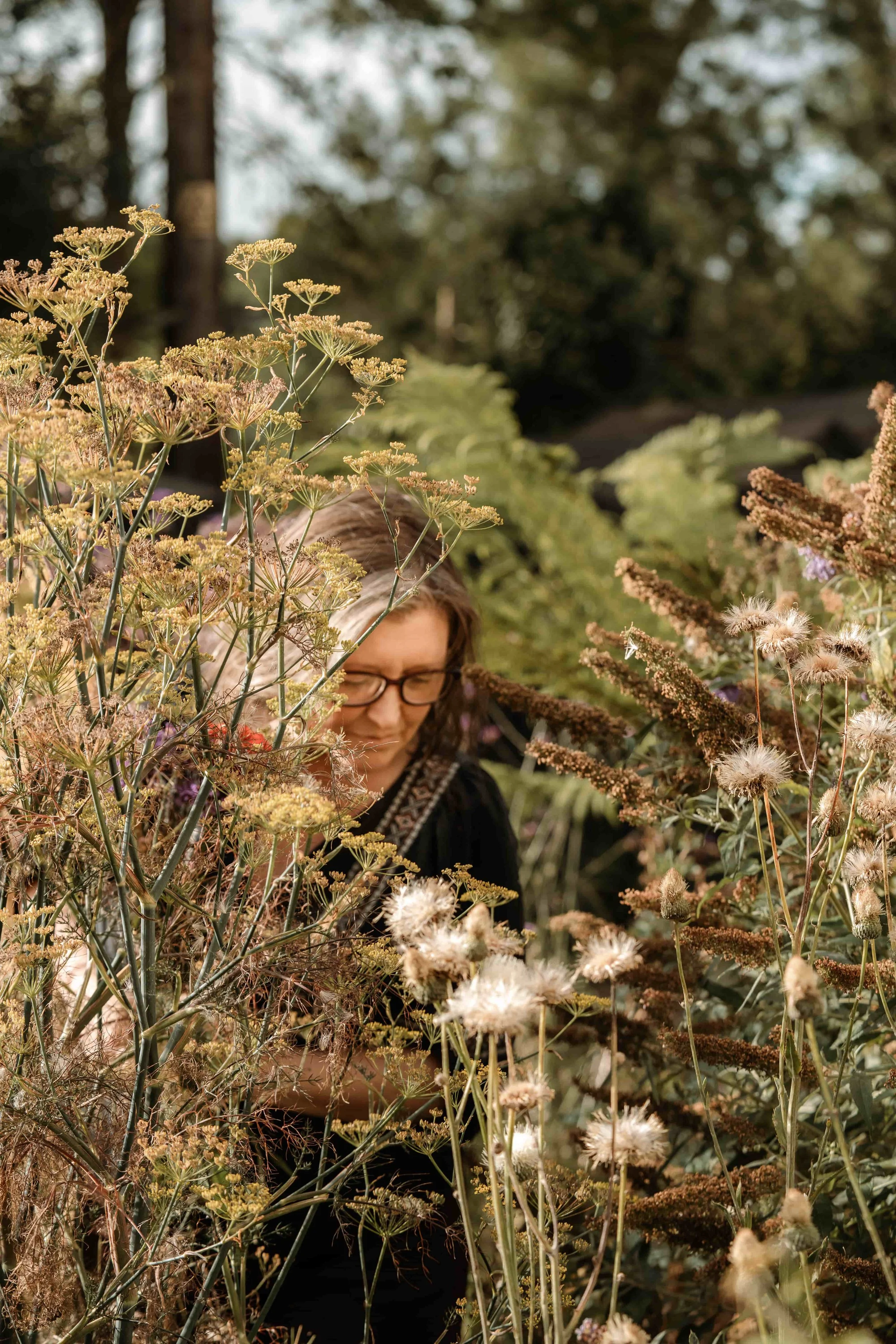 A woman with glasses and long hair surrounded by a dense tangle of various flowering plants and green foliage, outdoors in a natural setting.