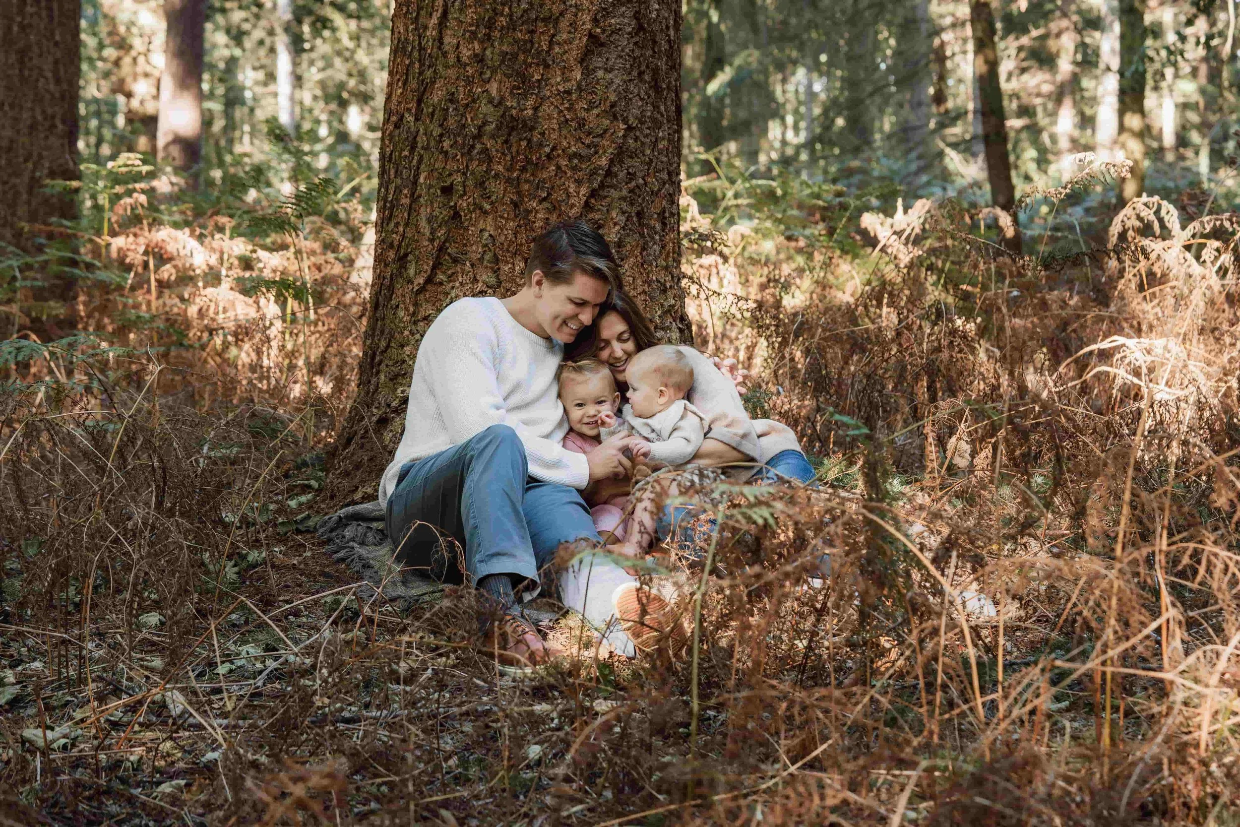 A family of four gathered at the base of a large tree in a forest, hugging and smiling, surrounded by fall foliage.