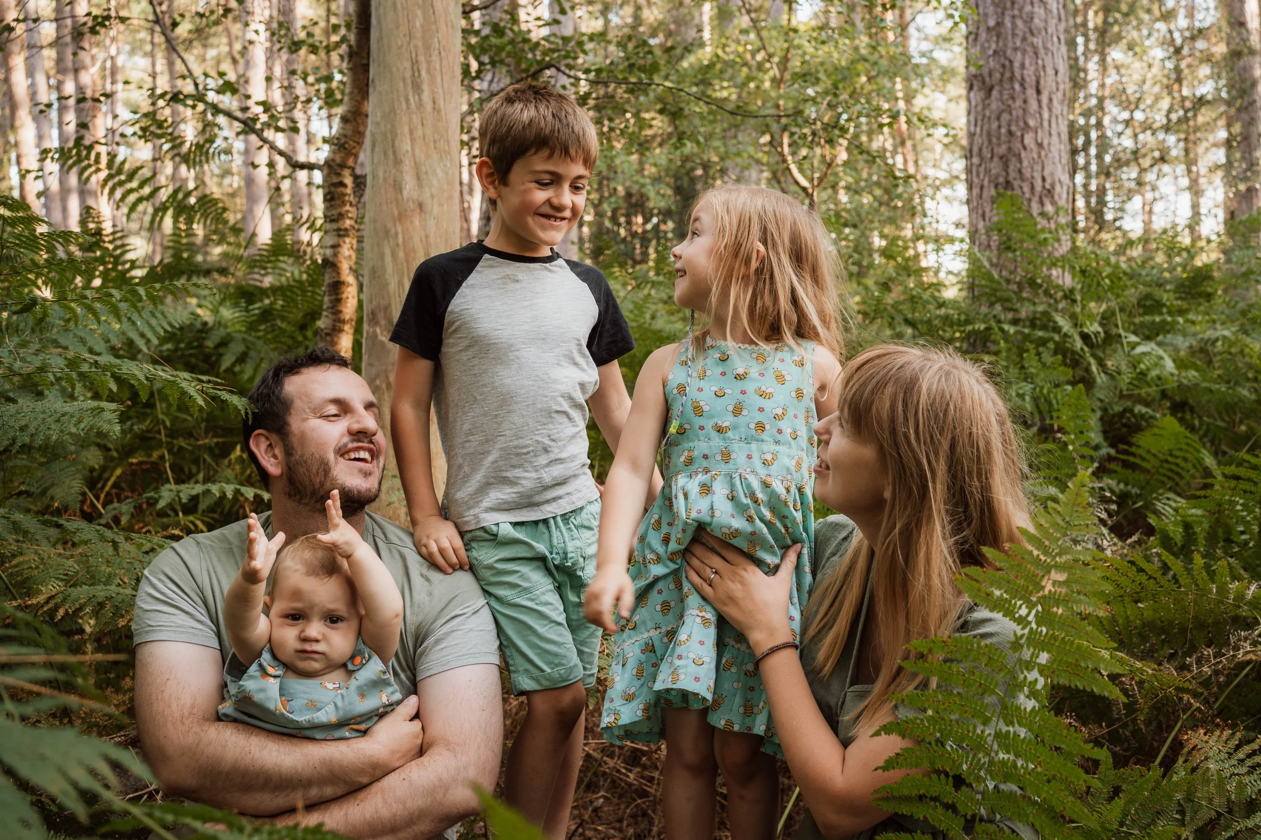 A family of six enjoying time outdoors in a forest, with two children standing and smiling at each other, a baby being held by a man, and a woman looking at the little girl.