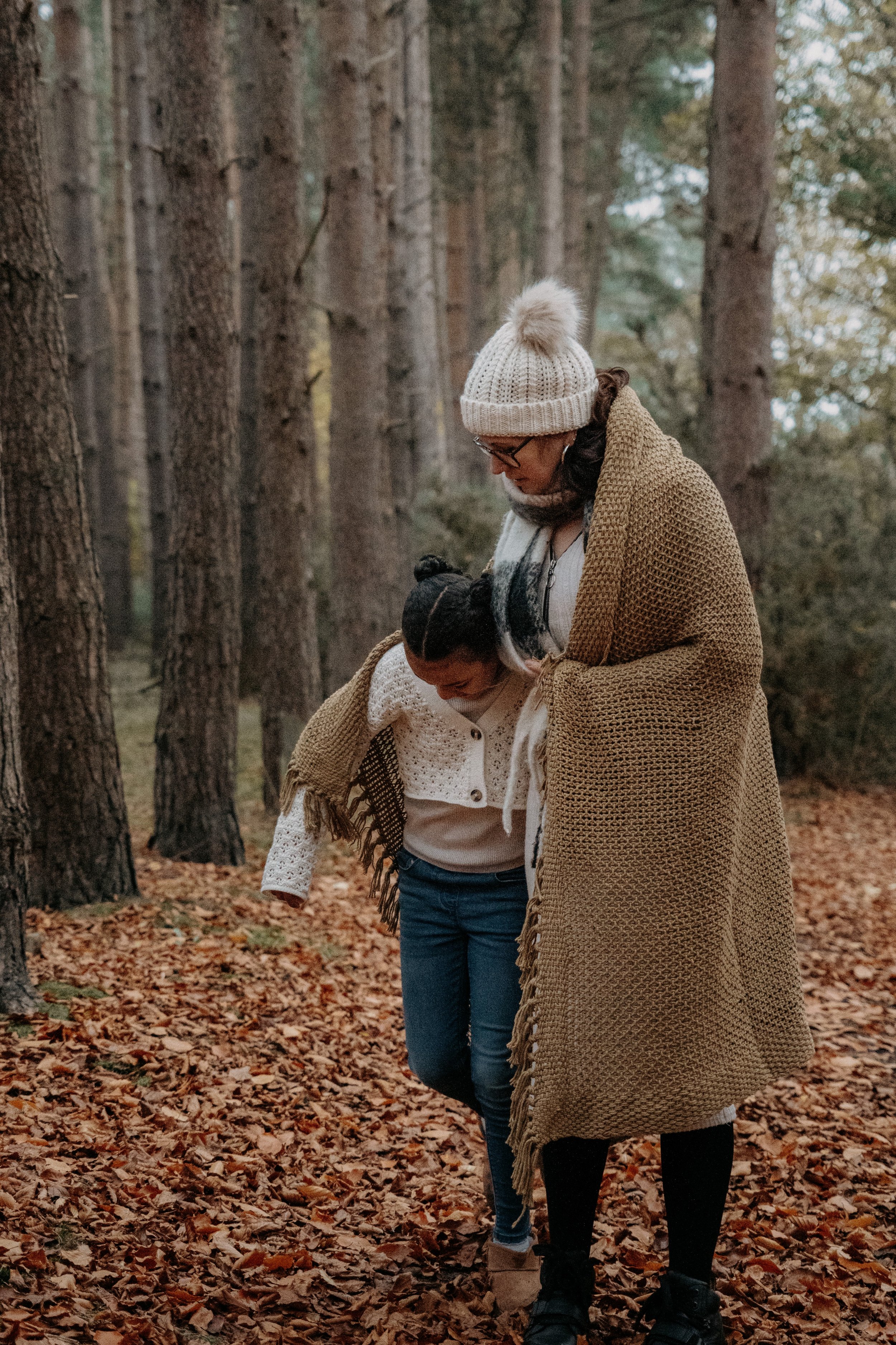 A woman and a girl walking together in a forest with autumn leaves on the ground, both dressed warmly for fall.