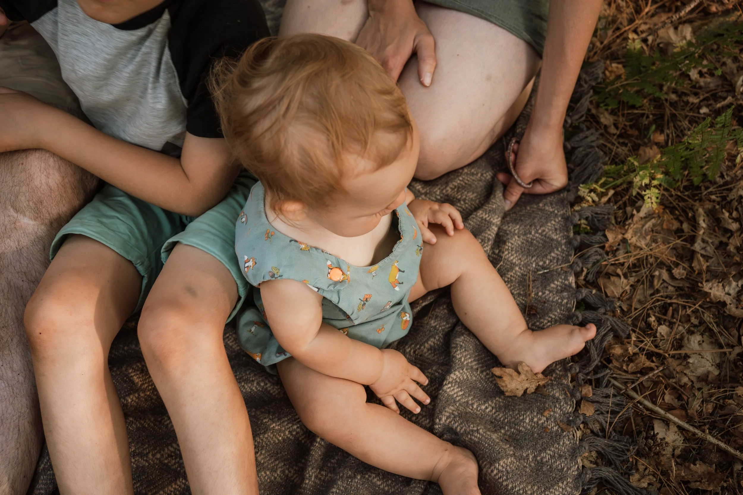 A young child with red hair, wearing a blue dress with animal prints, sitting on the lap of a person, surrounded by dried leaves and plants in an outdoor setting.