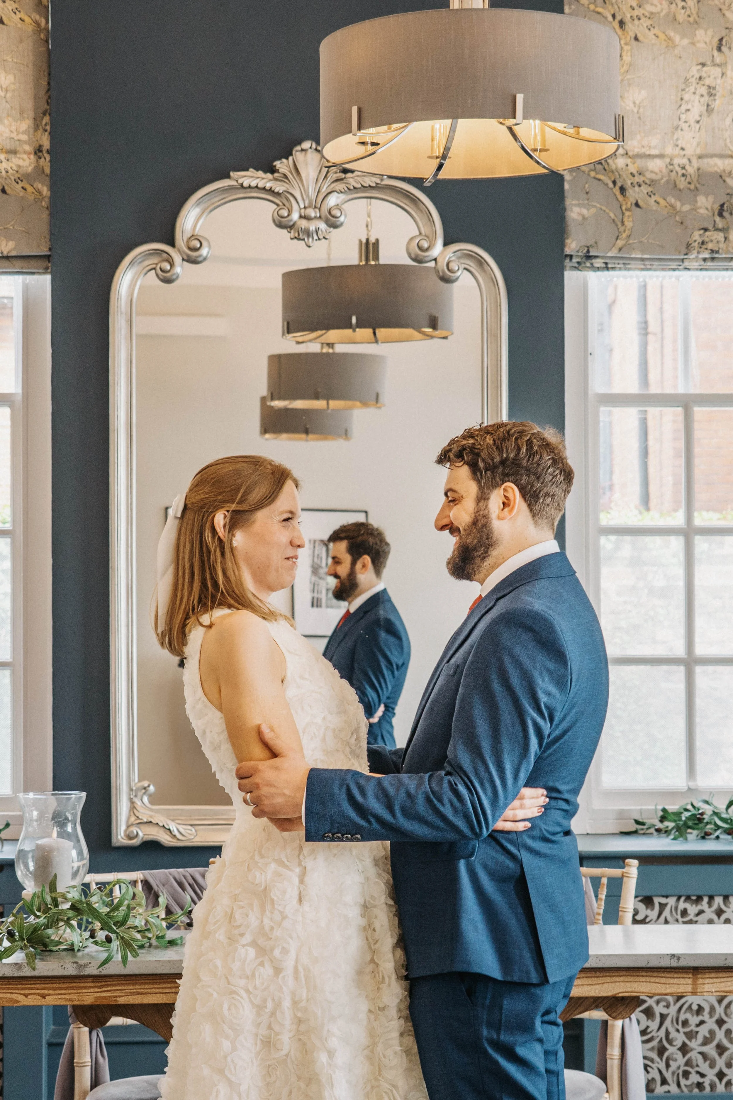 A bride and groom holding hands and looking at each other, standing in front of a large mirror, during their wedding ceremony in a decorated indoor space.