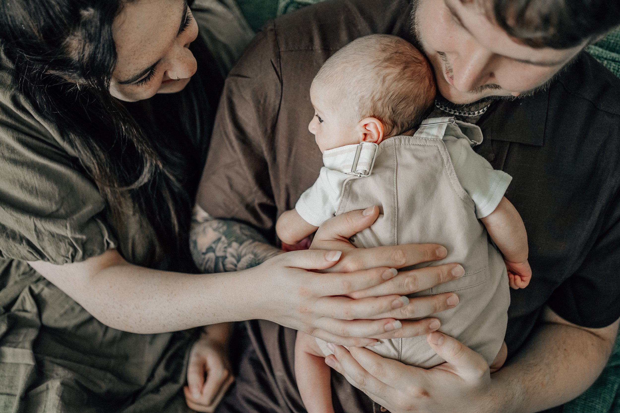 A baby being held by a man, with a woman touching the baby's back. The baby is resting on the man's chest, and the scene shows a close, tender moment among two adults and a child.