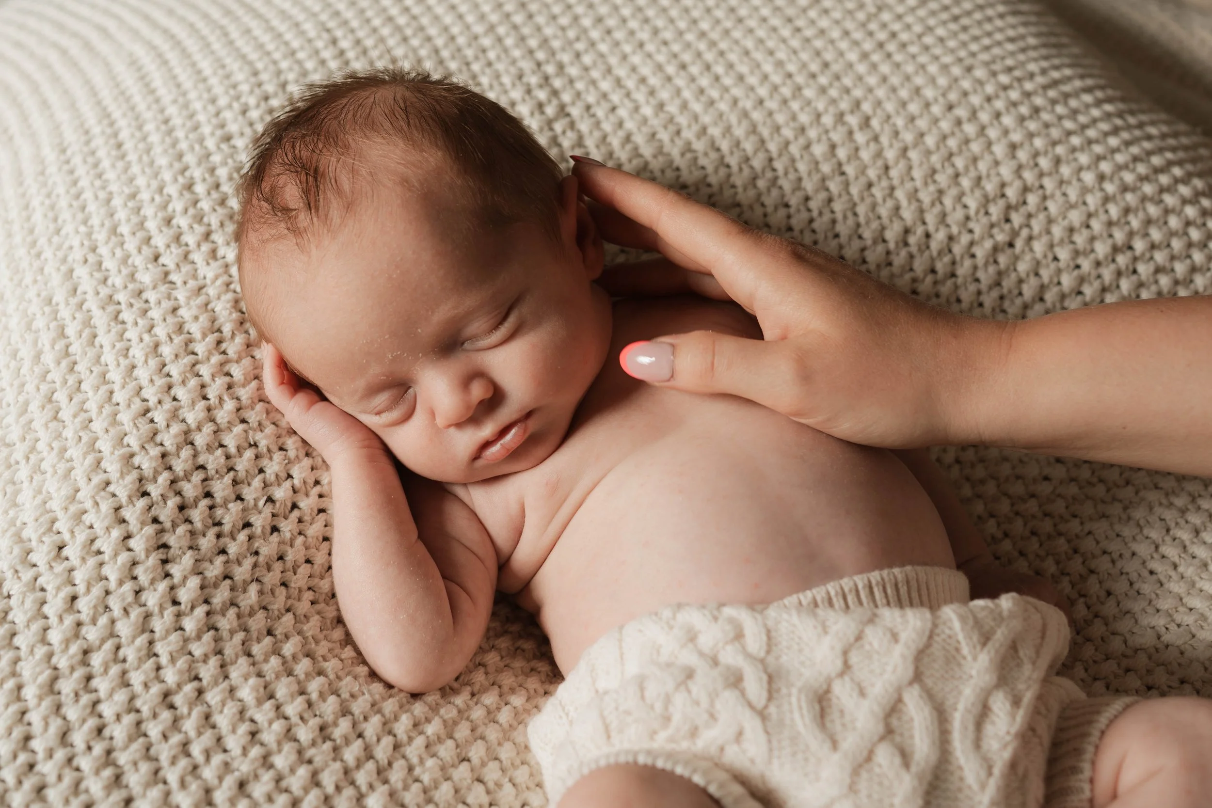 A sleeping baby resting on a textured beige blanket with a woman's hand gently touching their shoulder.