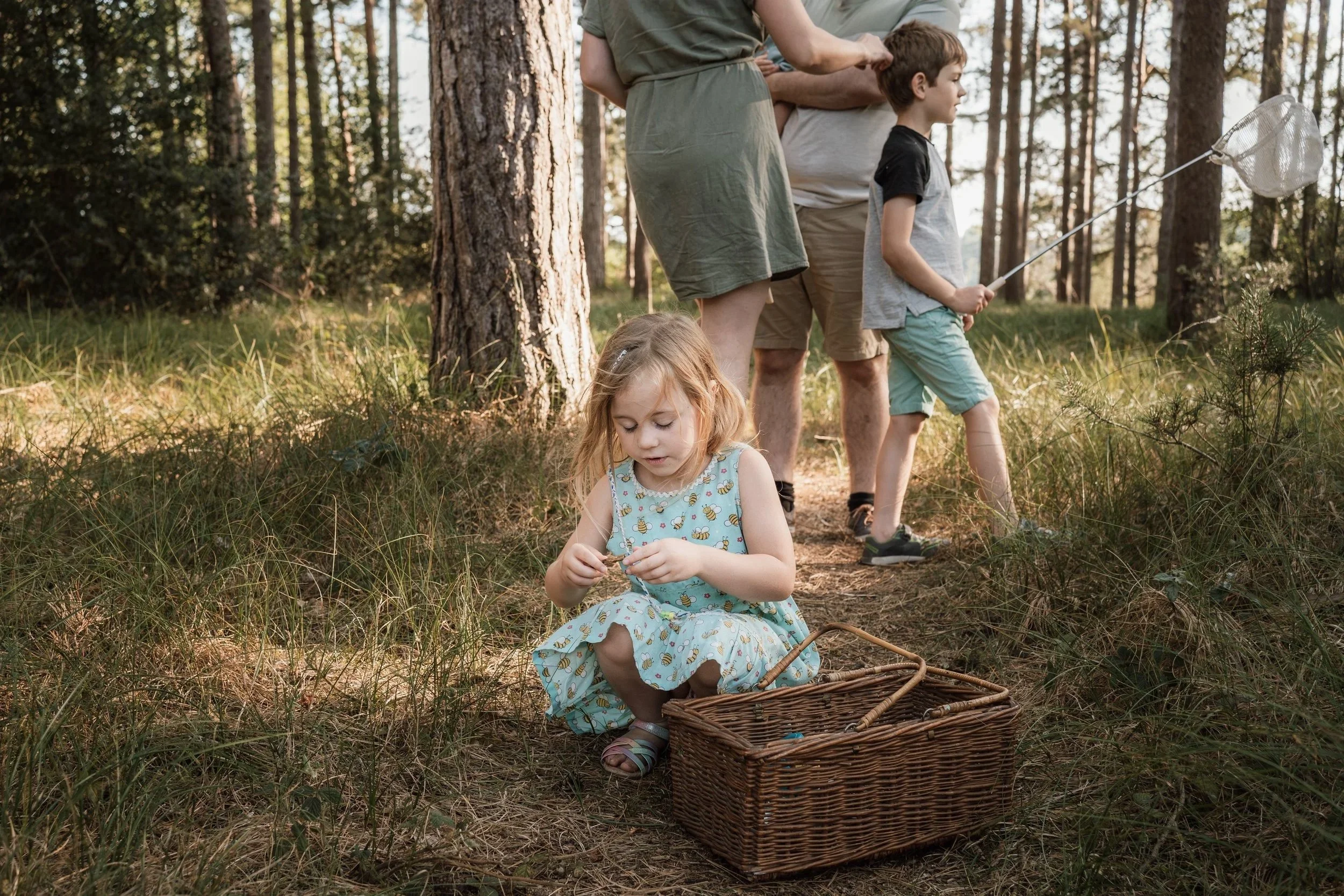 A young girl sitting on the ground in a forest, holding a small object, with a basket nearby. In the background, a boy and two adults are standing, one of whom is holding a butterfly net.