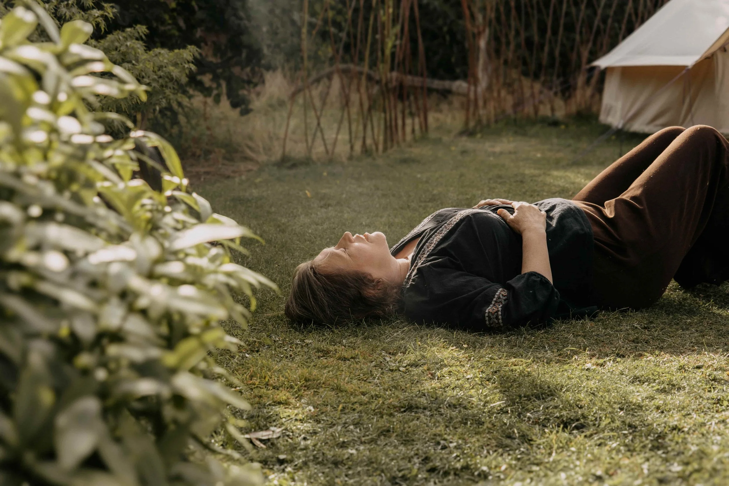 A woman lying on the grass near a tent in a wooded area, with greenery in the foreground and trees in the background, during dusk or evening.